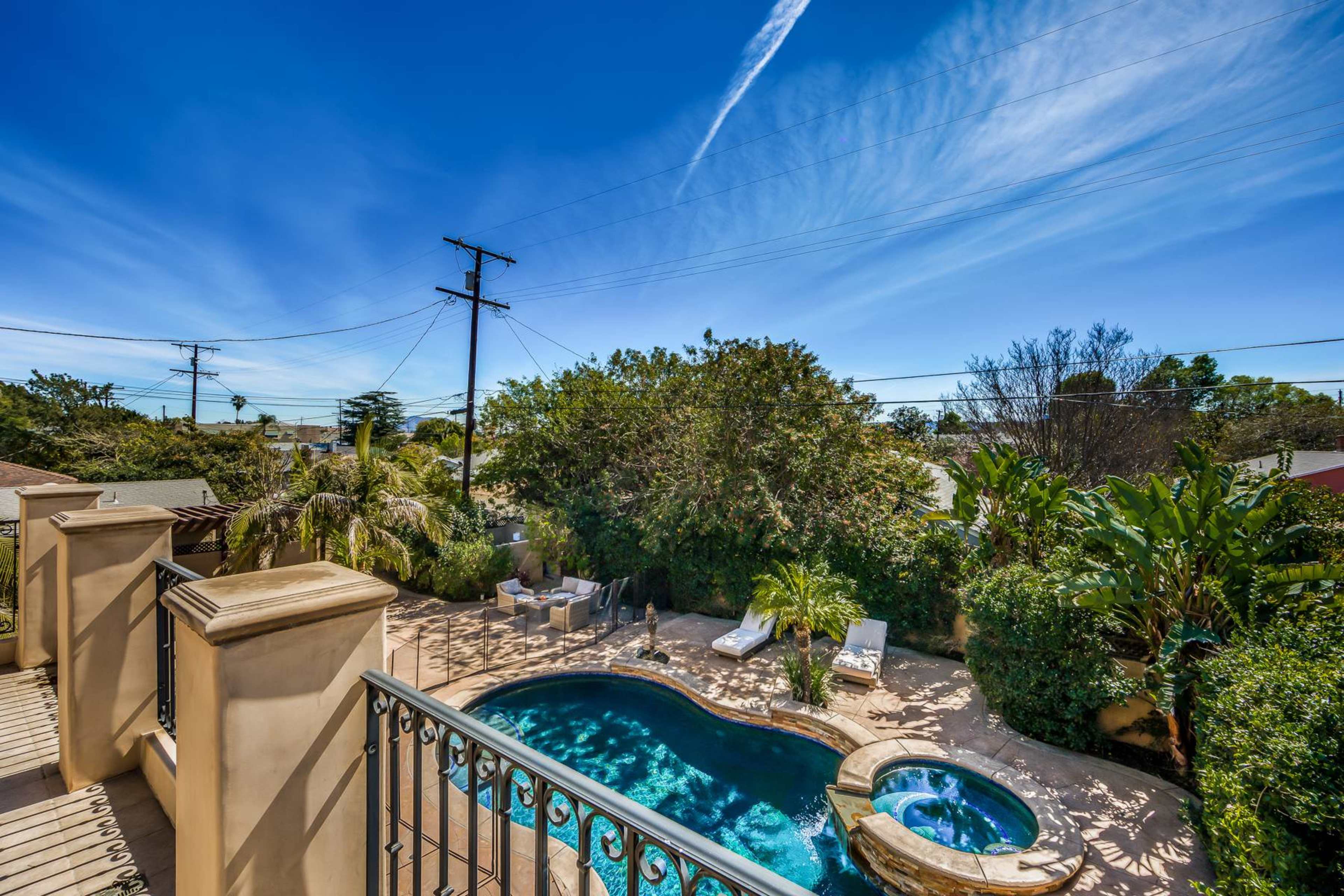 The image shows a backyard with a swimming pool, a jacuzzi, lounge chairs, and surrounding greenery under a clear blue sky.
