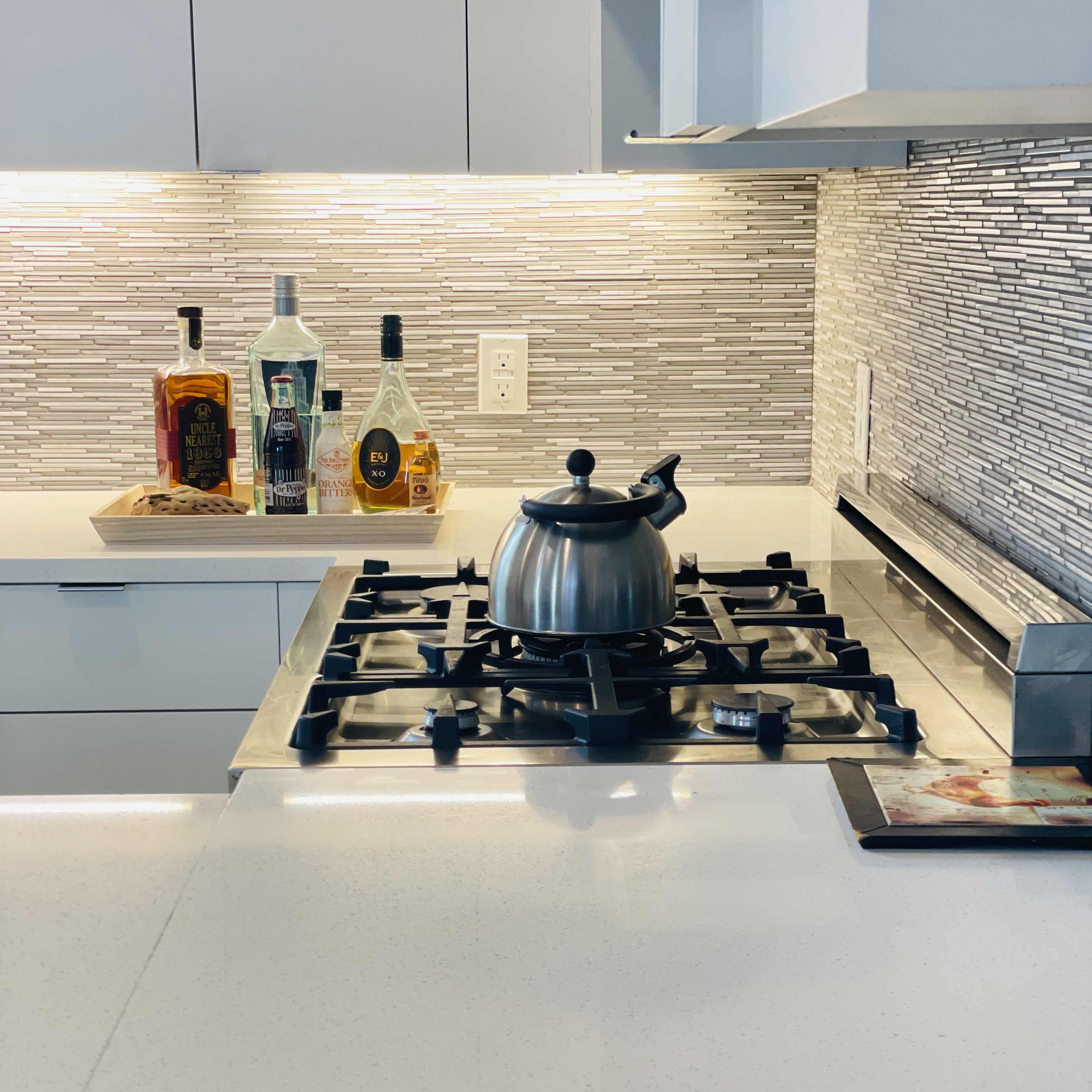 A stainless steel kettle sits on a black gas stove in a modern kitchen with a tiled backsplash and various bottles displayed on a tray nearby.