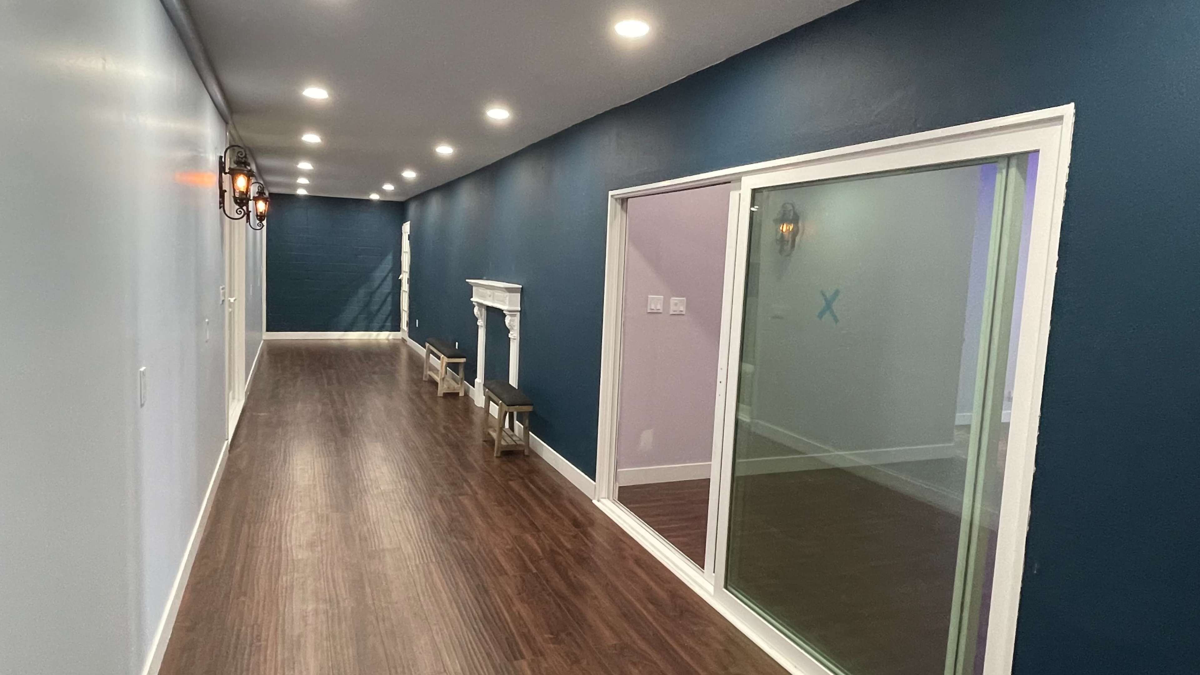 A long, well-lit hallway features blue and white walls, a wooden floor, and a glass door leading to a room with a fireplace and decorative benches along one side.