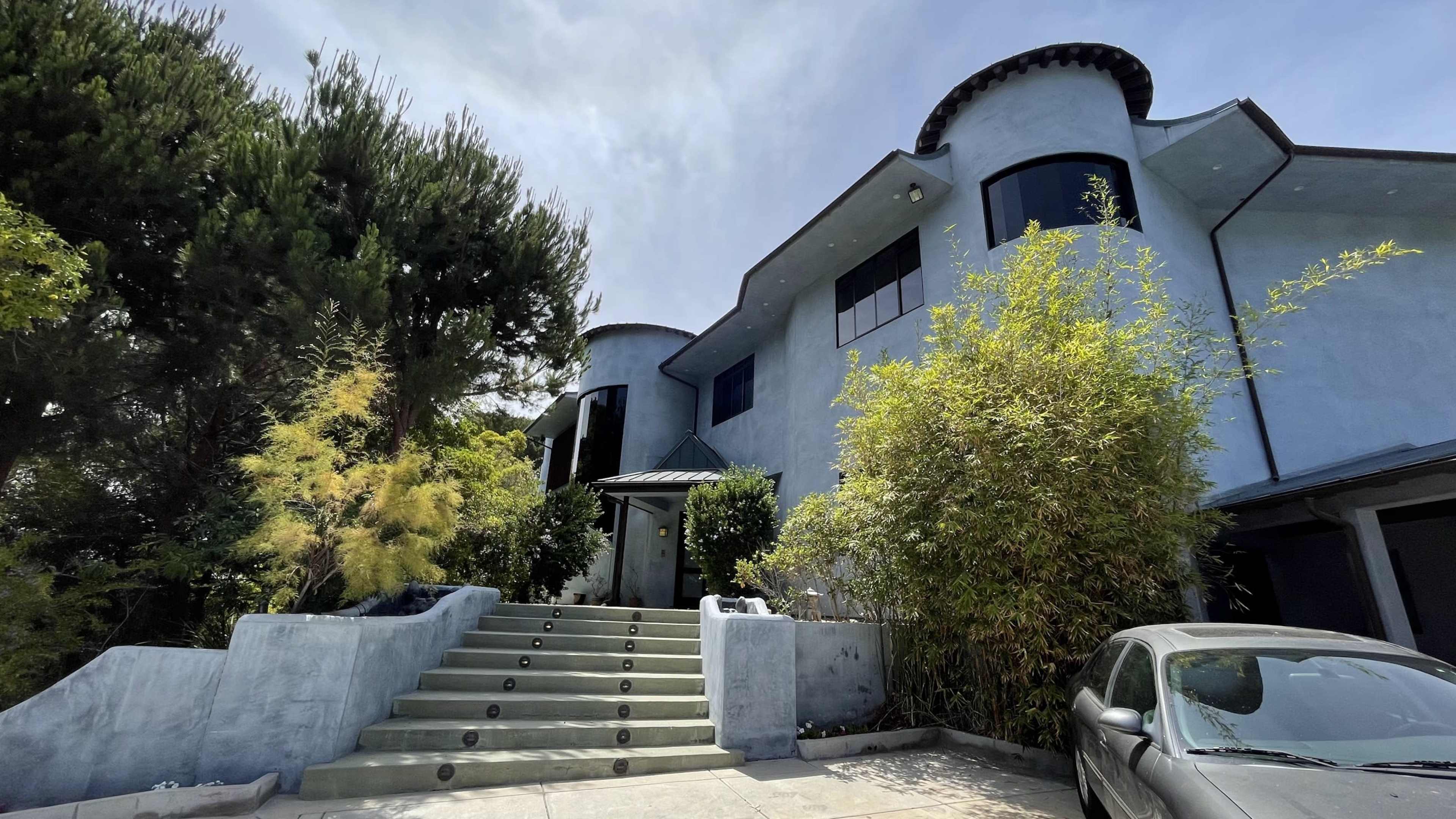 A modern blue home with a curved design and a stone staircase leads to the entrance, surrounded by trees and greenery.