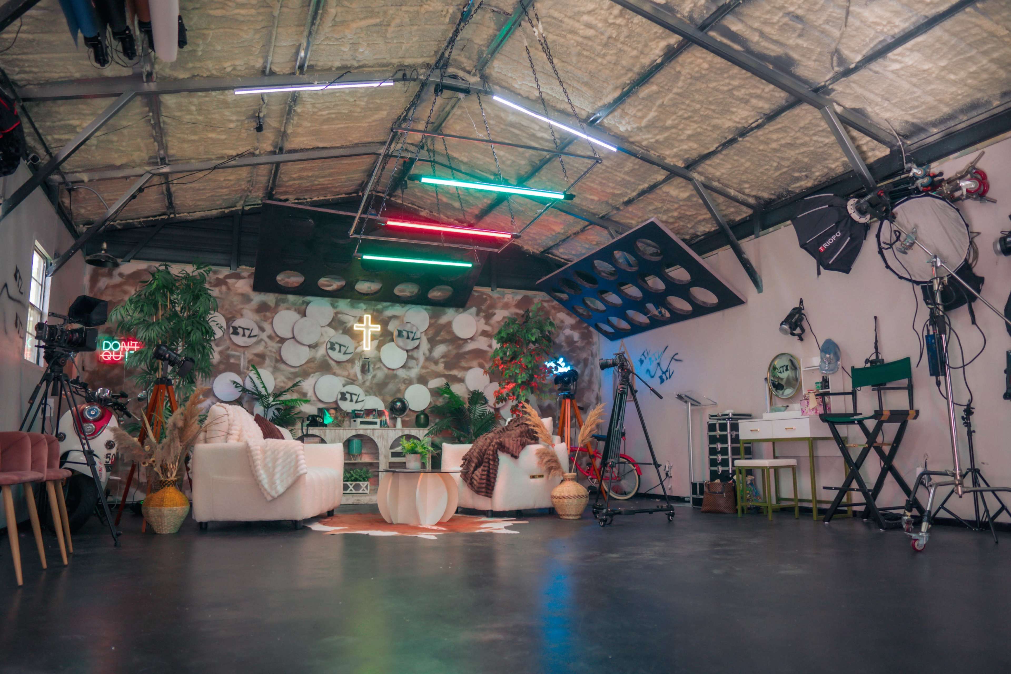 The image shows a spacious, brightly lit studio interior featuring two white sofas, a circular wooden coffee table, various lighting equipment, and decorative plants.