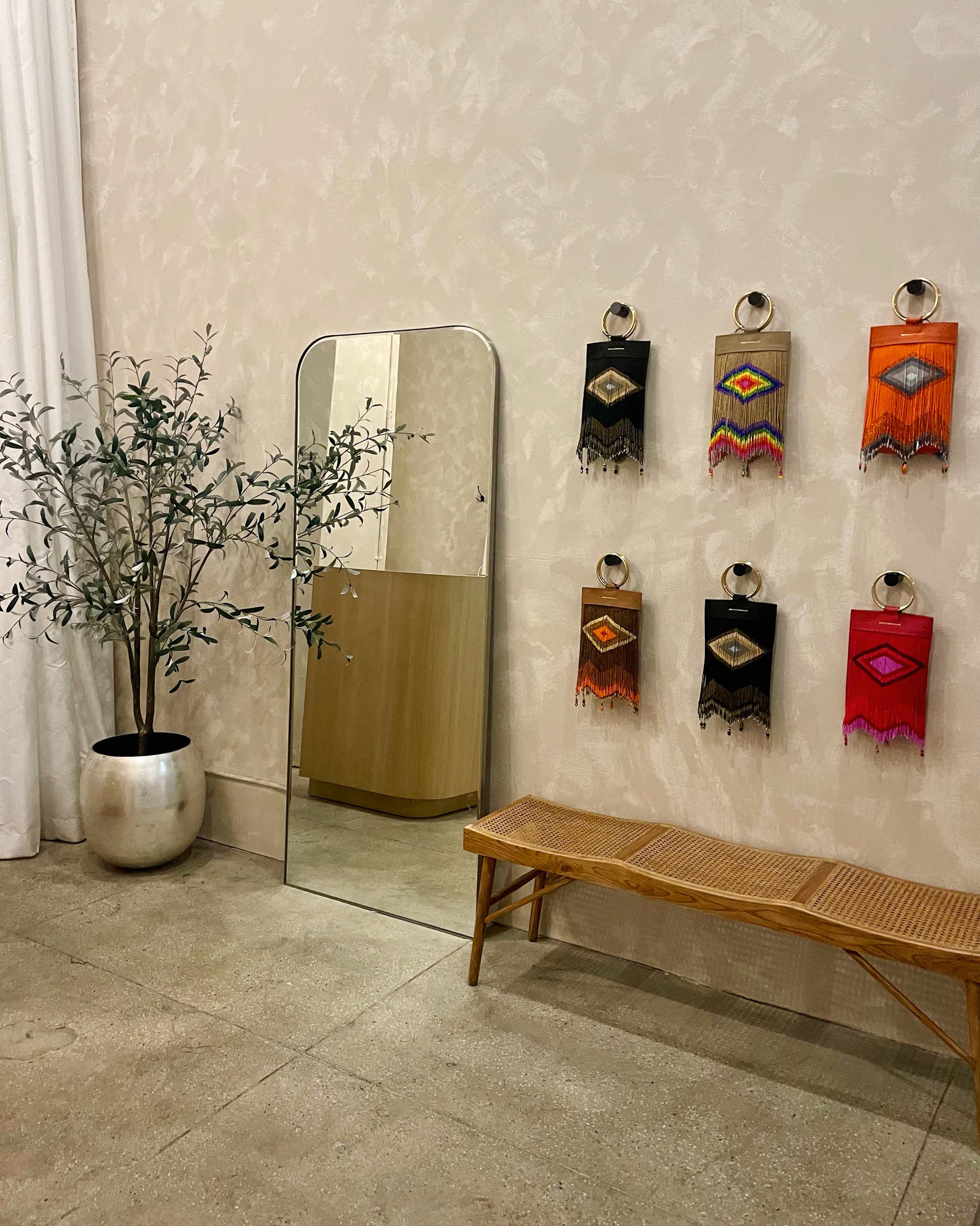 A minimalist interior featuring a tall mirror, a decorative plant in a silver pot, a wooden bench, and six colorful woven wall hangings.
