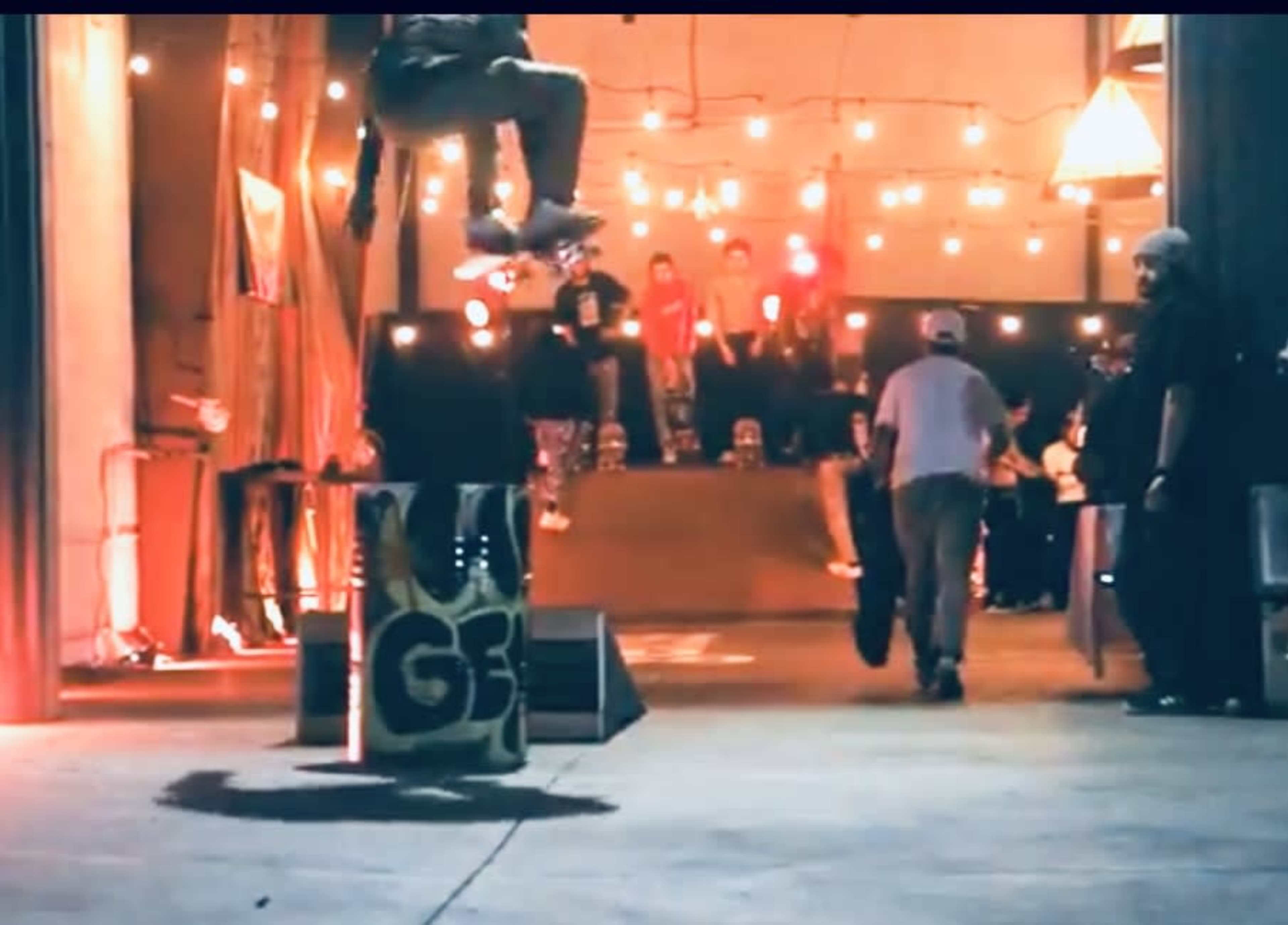 A skateboarder performs a trick in mid-air above a large container while spectators watch from a raised platform in a well-lit indoor space.
