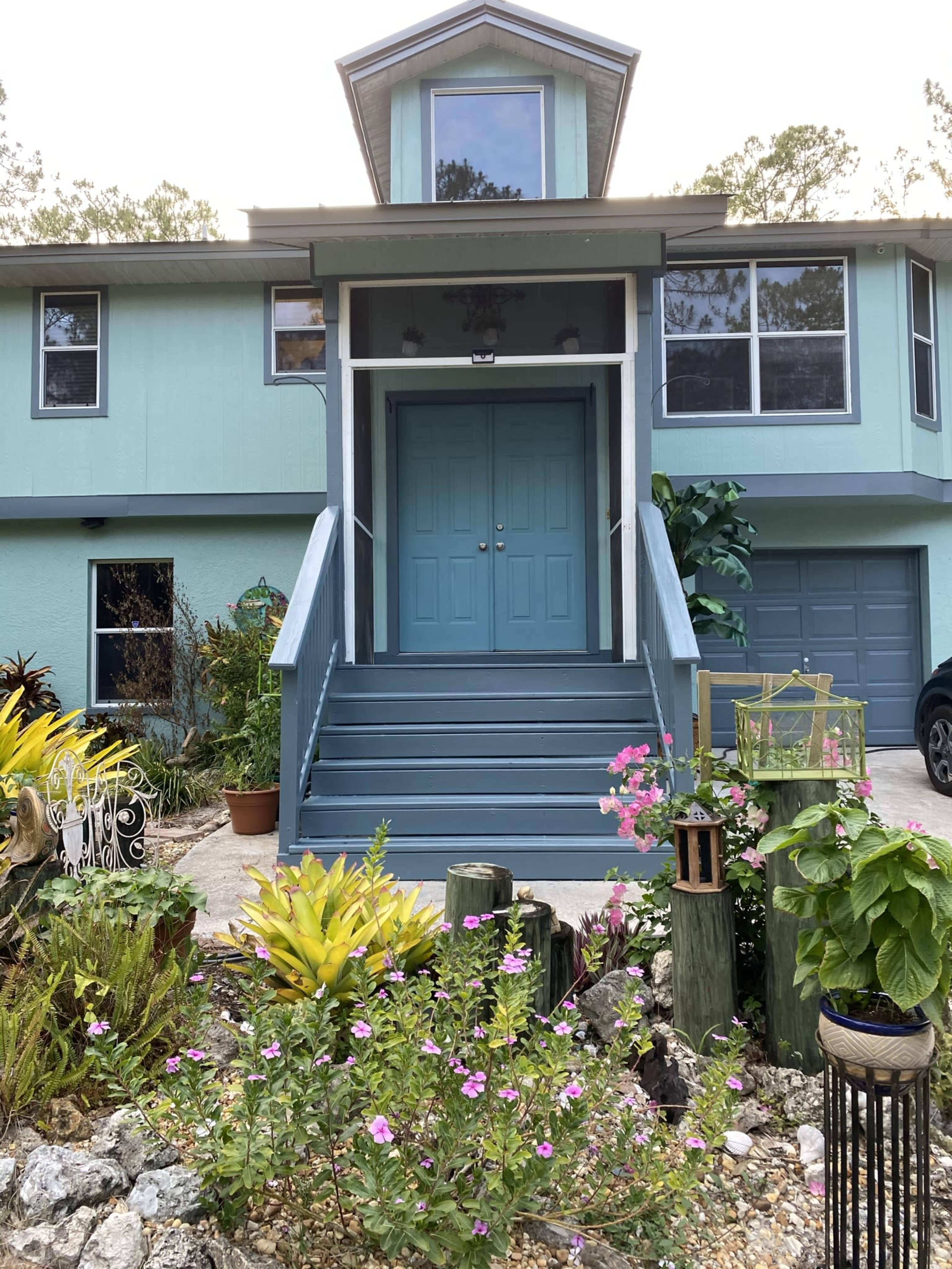 A two-story house with a light blue exterior features a double front door and a landscaped garden with colorful plants and flowers.