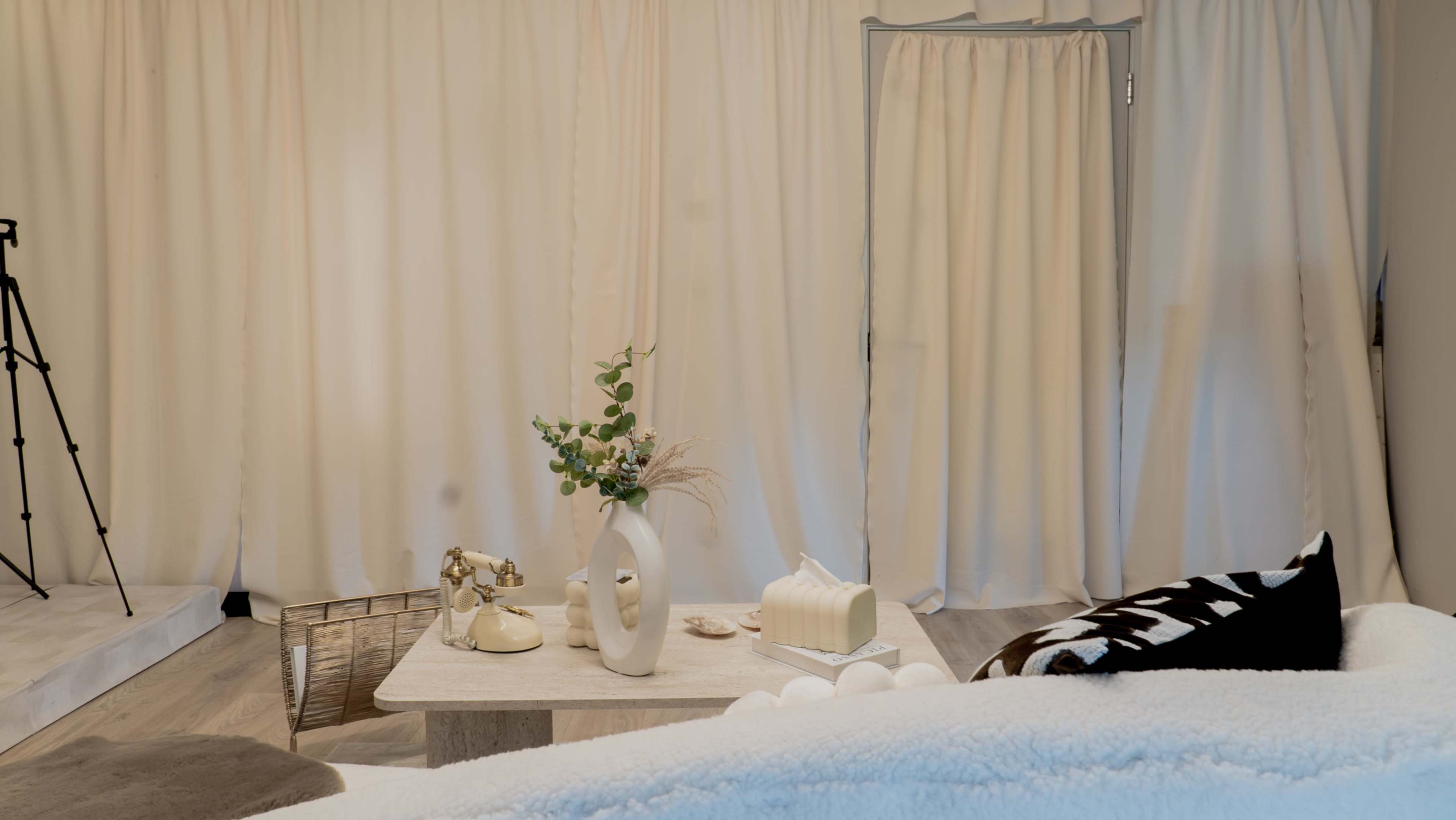 The image shows a softly lit room featuring a table with decorative items, a vase of greenery, and a curtain separating the space.