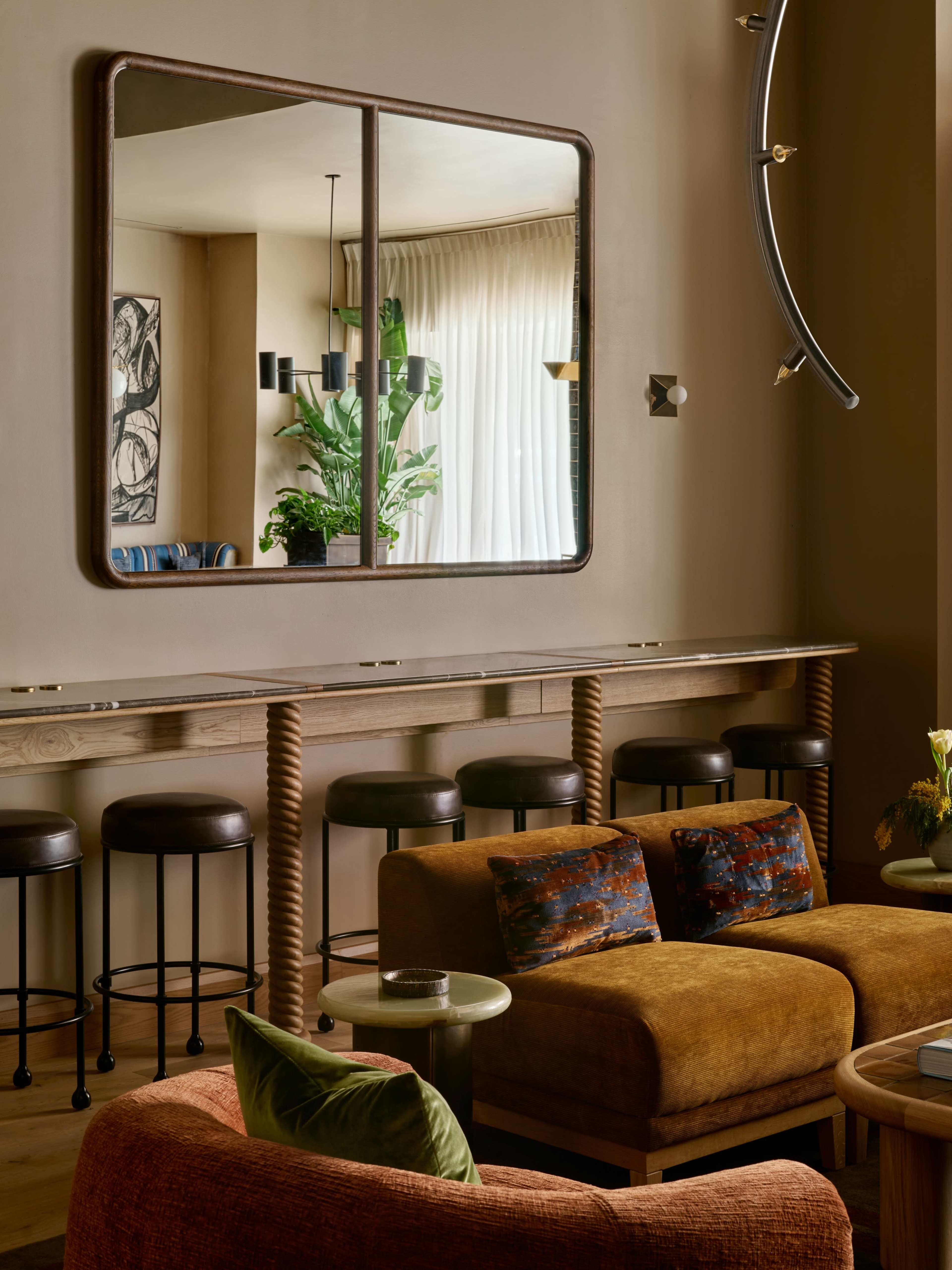 The image shows a stylish interior featuring a wooden bar with stools, a large mirror on the wall, and a seating area with two sofas and decorative cushions.