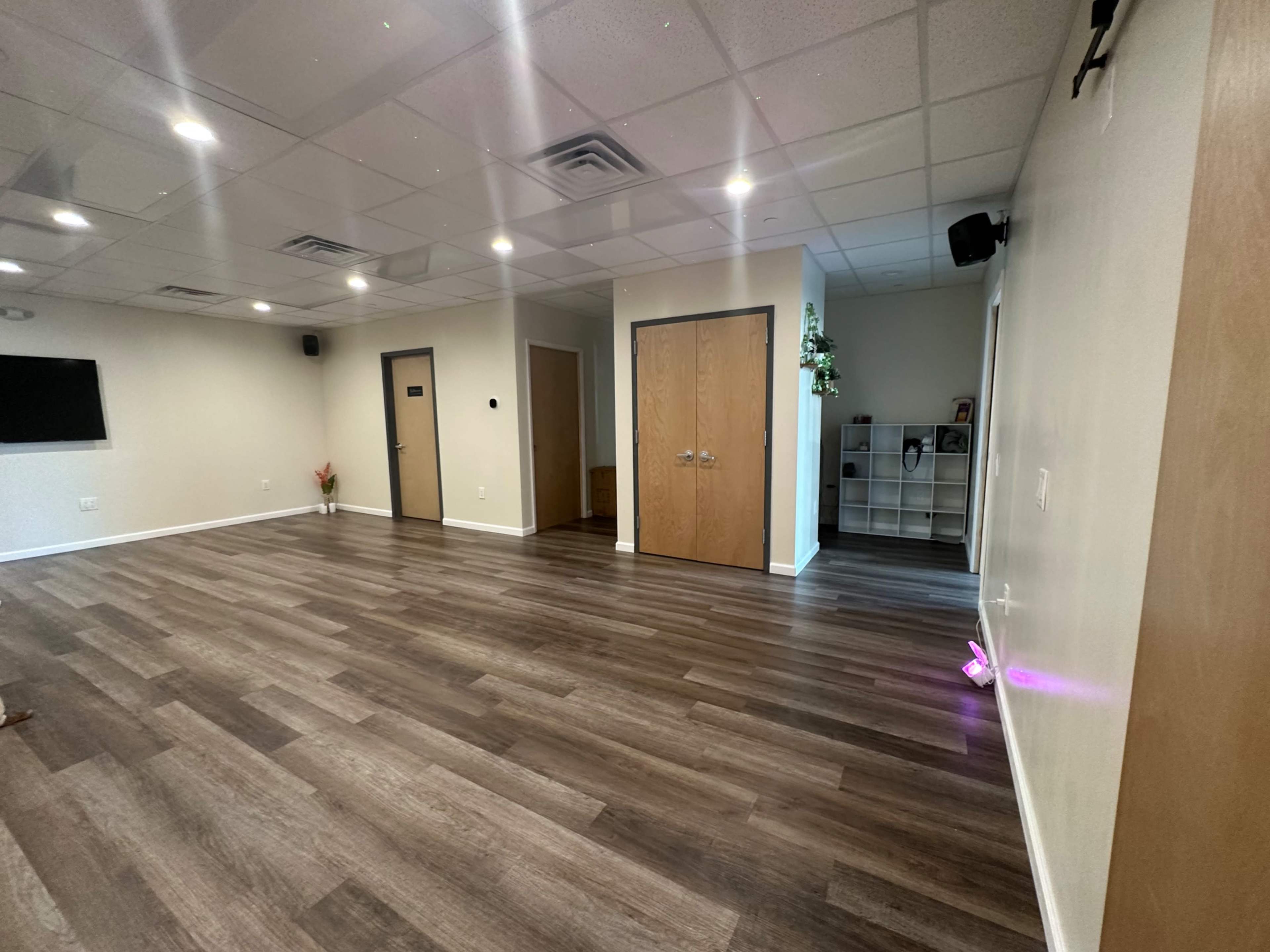 A spacious room with wooden flooring, a few doors, and minimal furnishings, including a wall-mounted TV on one side.