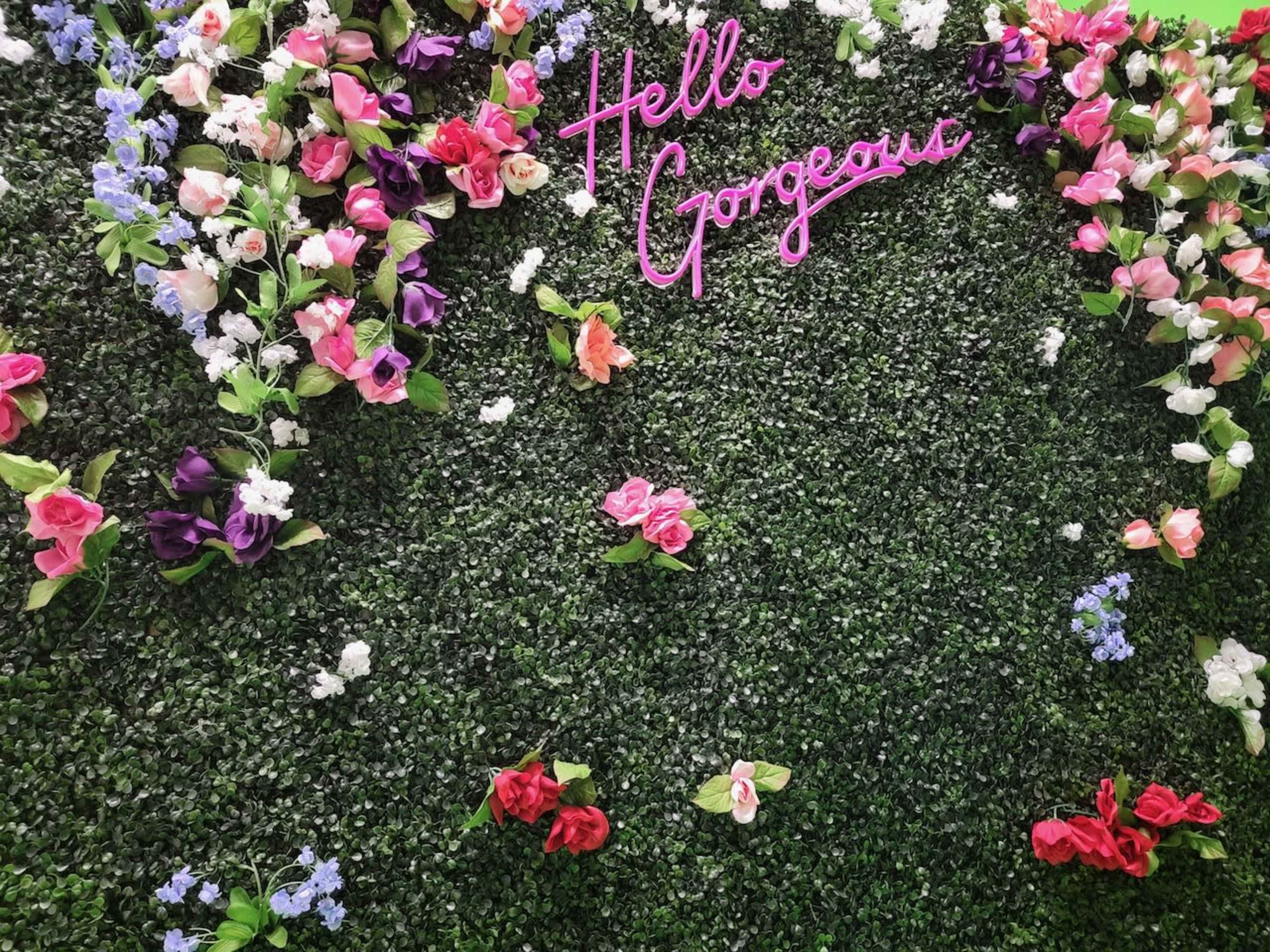 A green floral backdrop features a pink neon sign that reads "Hello Gorgeous" surrounded by various artificial flowers.