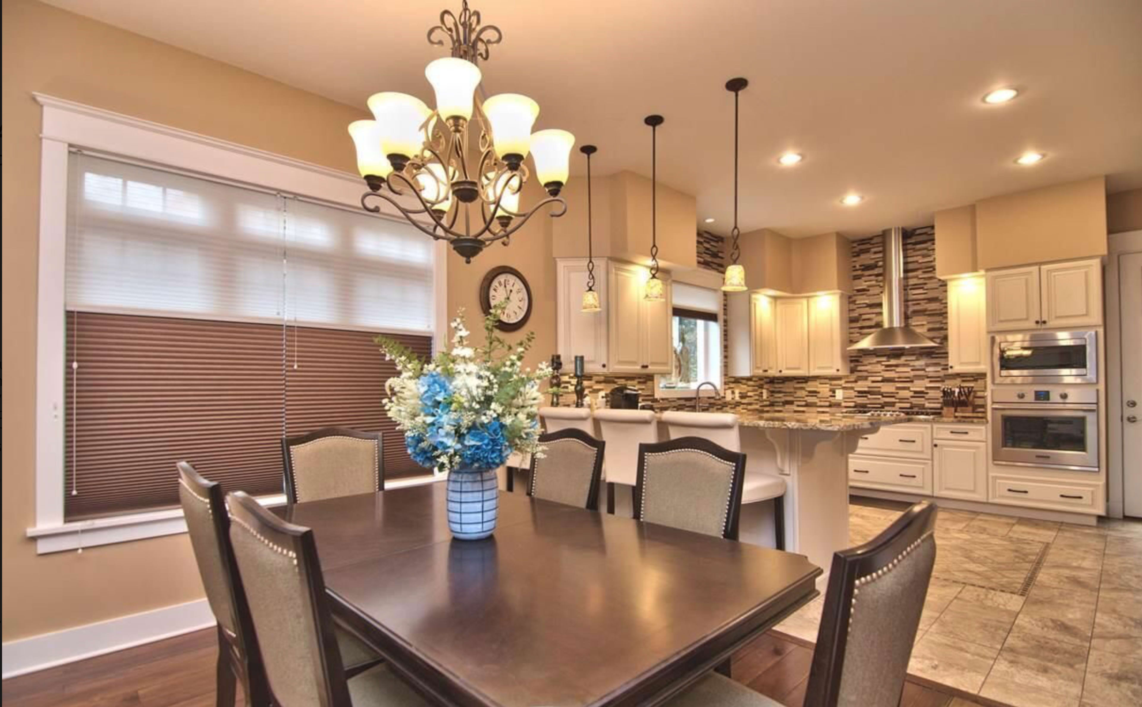 A dining area features a wooden table surrounded by chairs, with a chandelier above and a modern kitchen visible in the background.