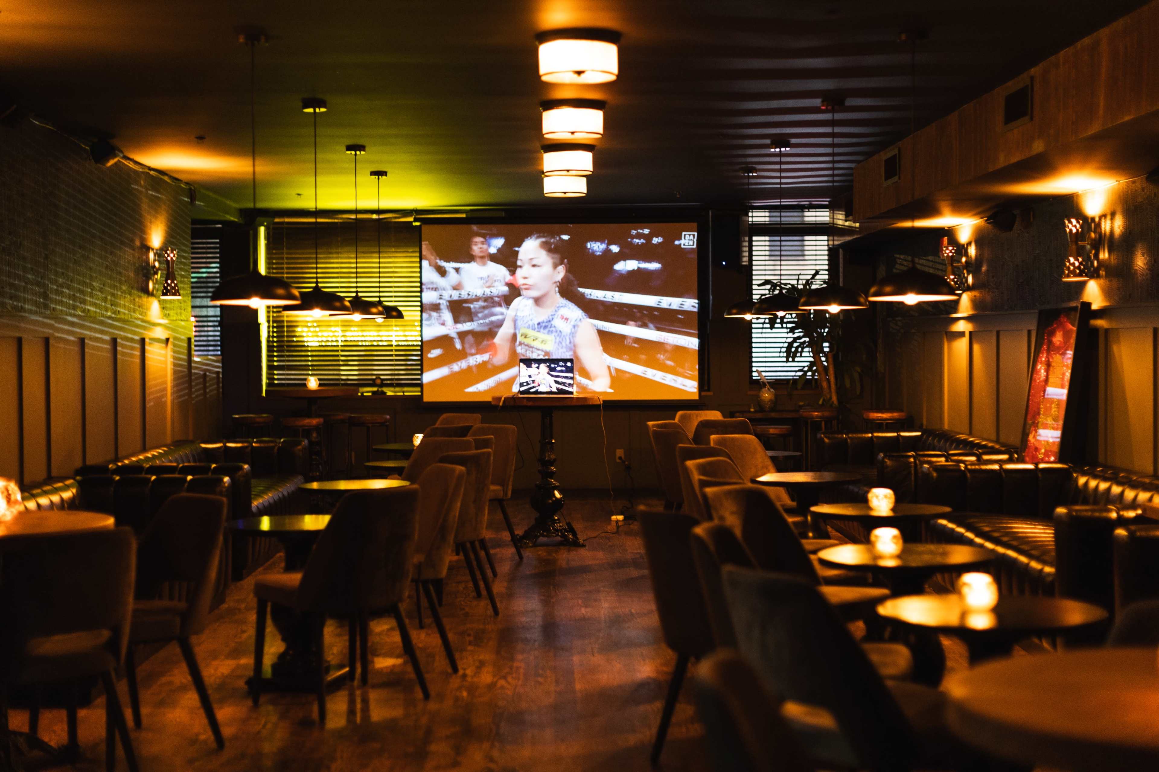 A dimly lit lounge features a large screen displaying a boxing match, with empty tables and chairs arranged around the room.