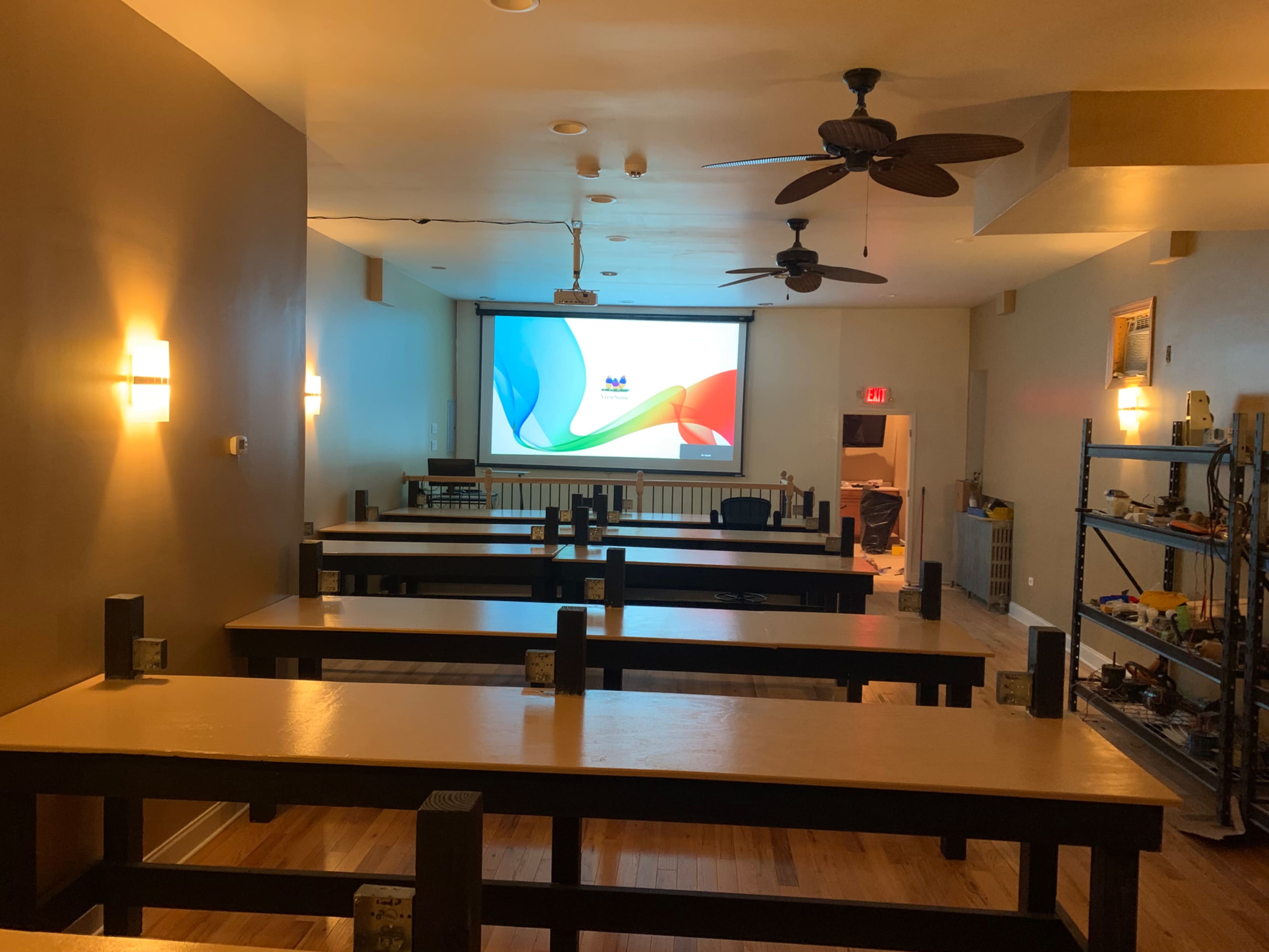 An empty room with several long tables, a projector screen on the front wall, and a shelf with various items in the corner.