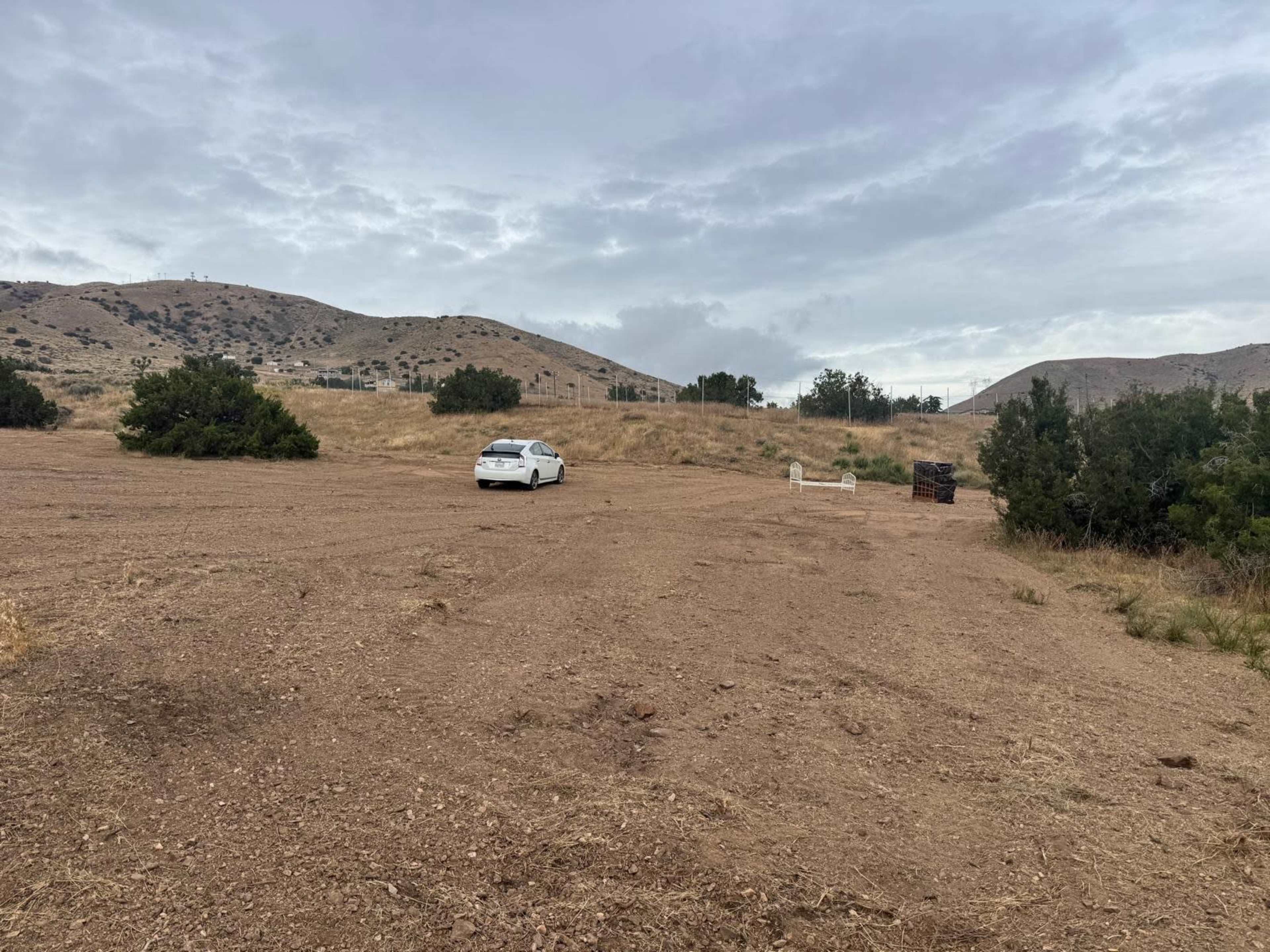 A white car is parked on a barren, sandy area surrounded by sparse vegetation and rolling hills under a cloudy sky.