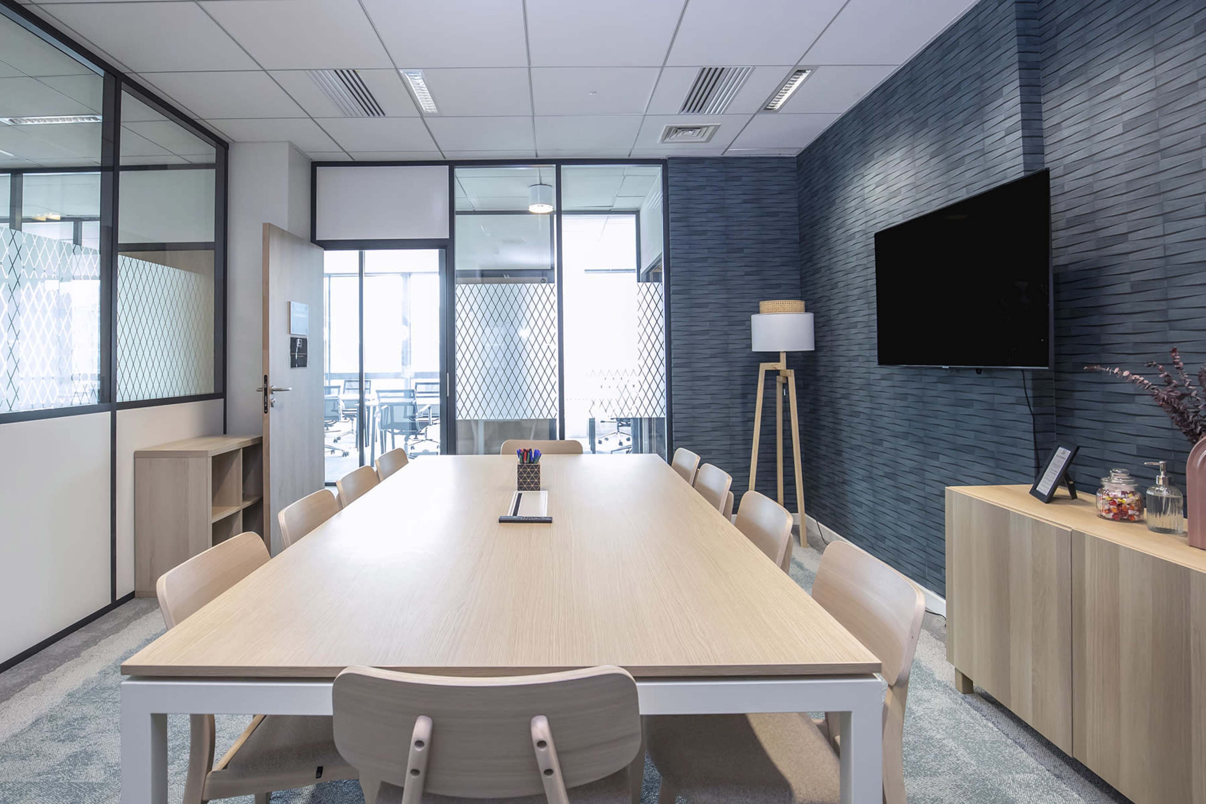 A modern meeting room with a large wooden table surrounded by chairs, a wall-mounted television, and glass partitioning allowing for natural light.