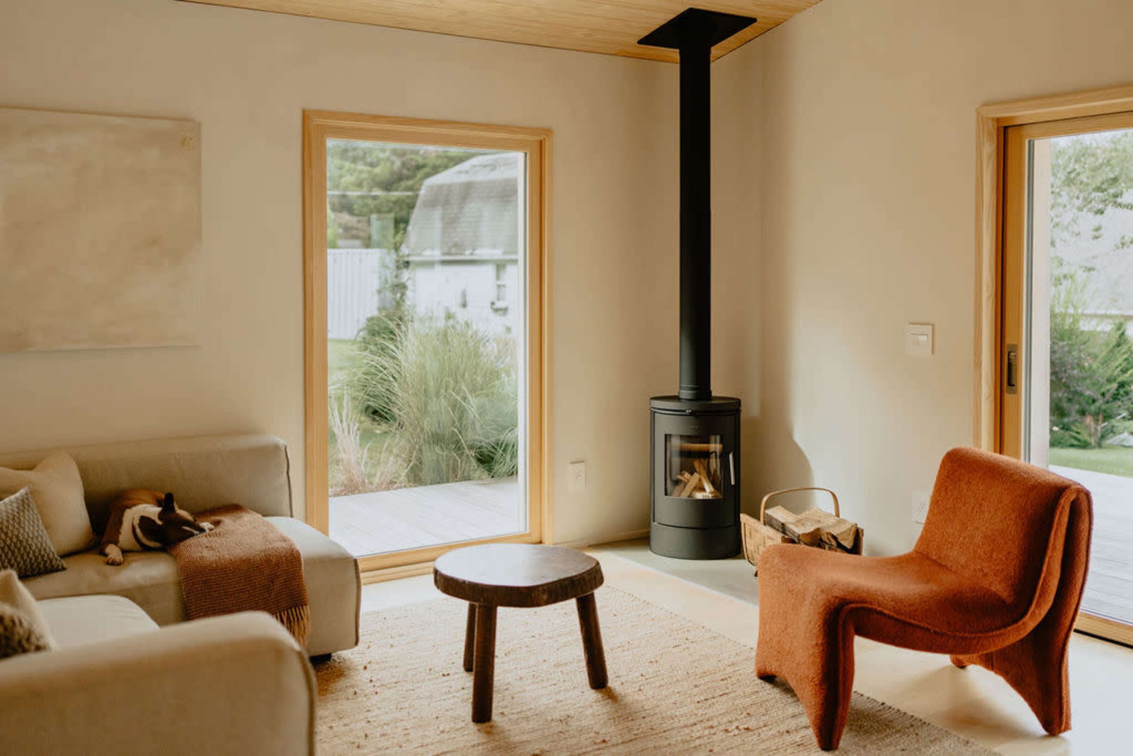 The scene depicts a cozy living room featuring a sofa, a round wooden coffee table, a modern wood stove, and large windows that open to a view of greenery outside.