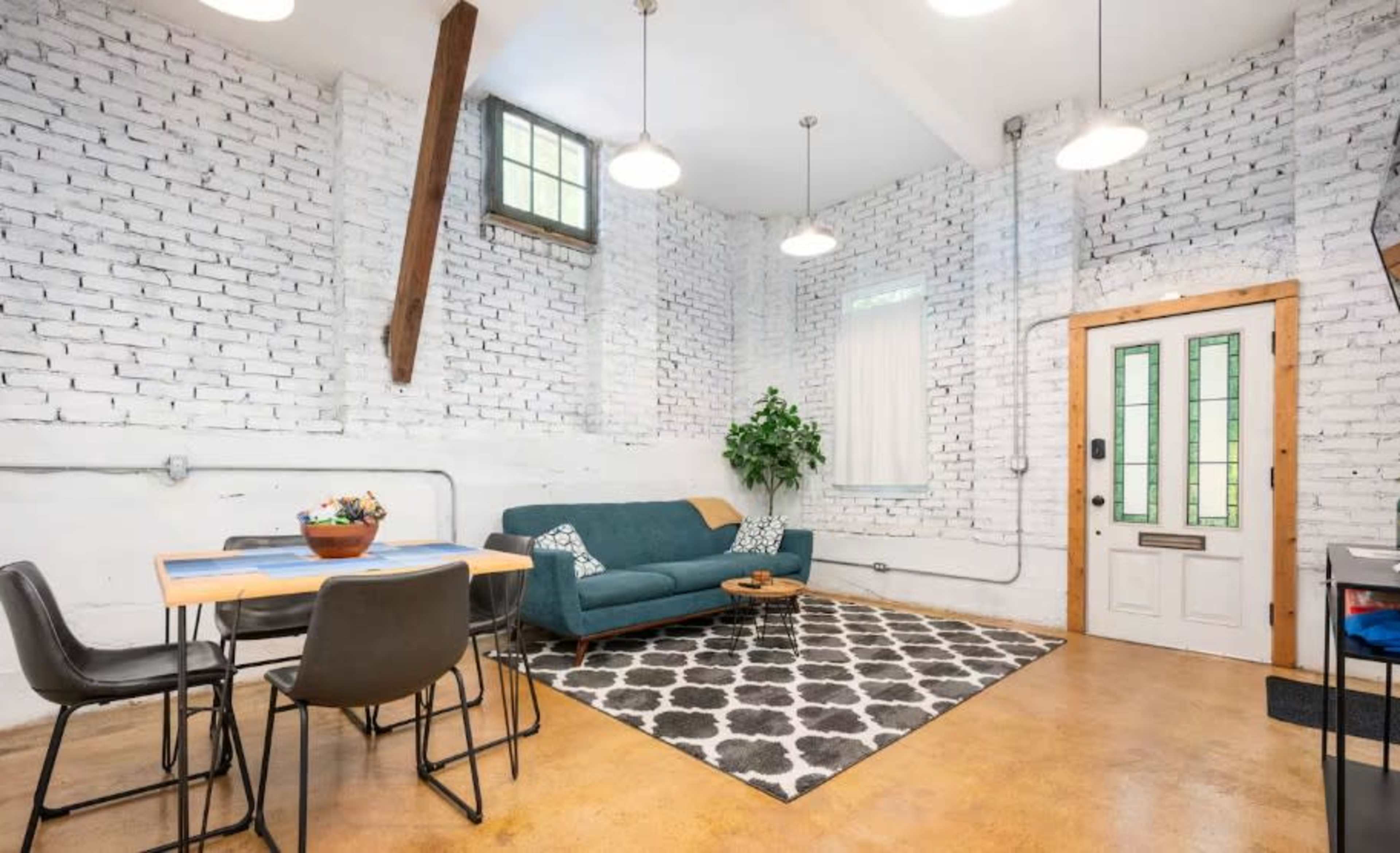 The image shows a bright, loft-style living area with white brick walls, a teal sofa, a dining table with black chairs, and a patterned rug.