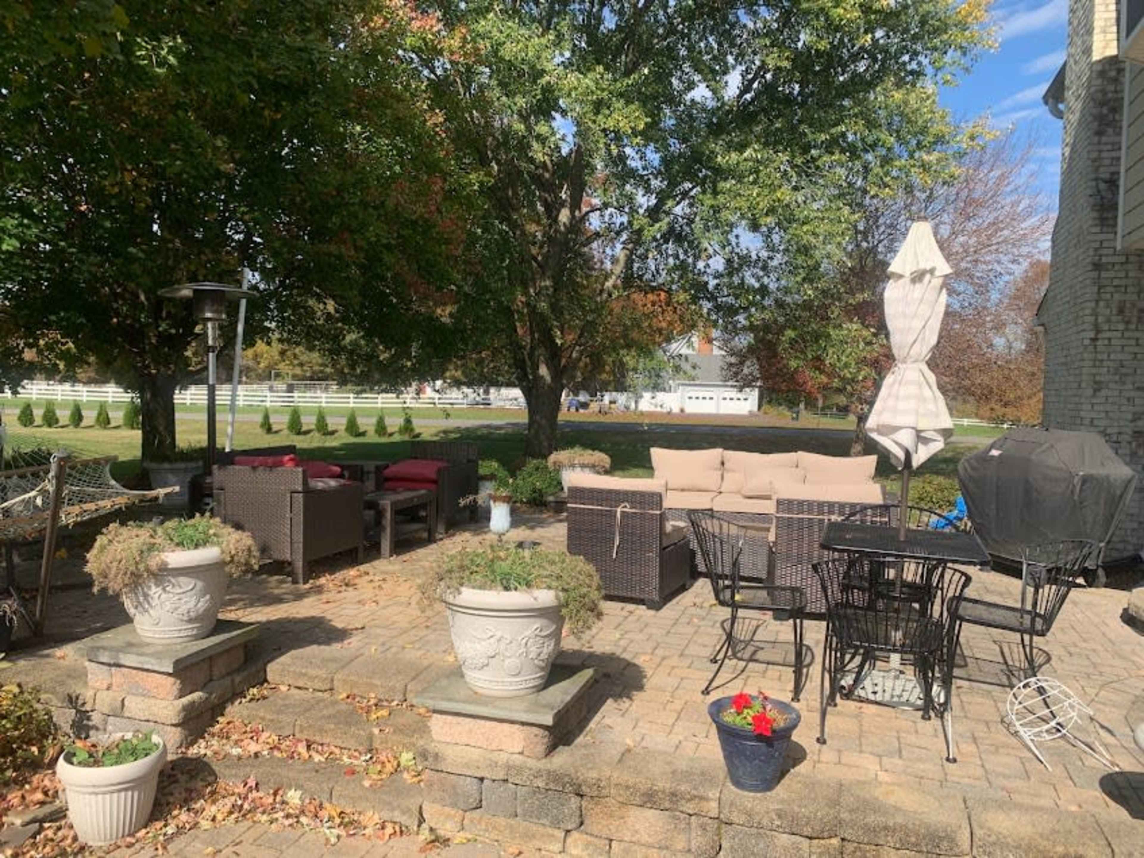 A patio area with wicker furniture, a table and chairs, potted plants, and a grill, surrounded by trees and grassy space.