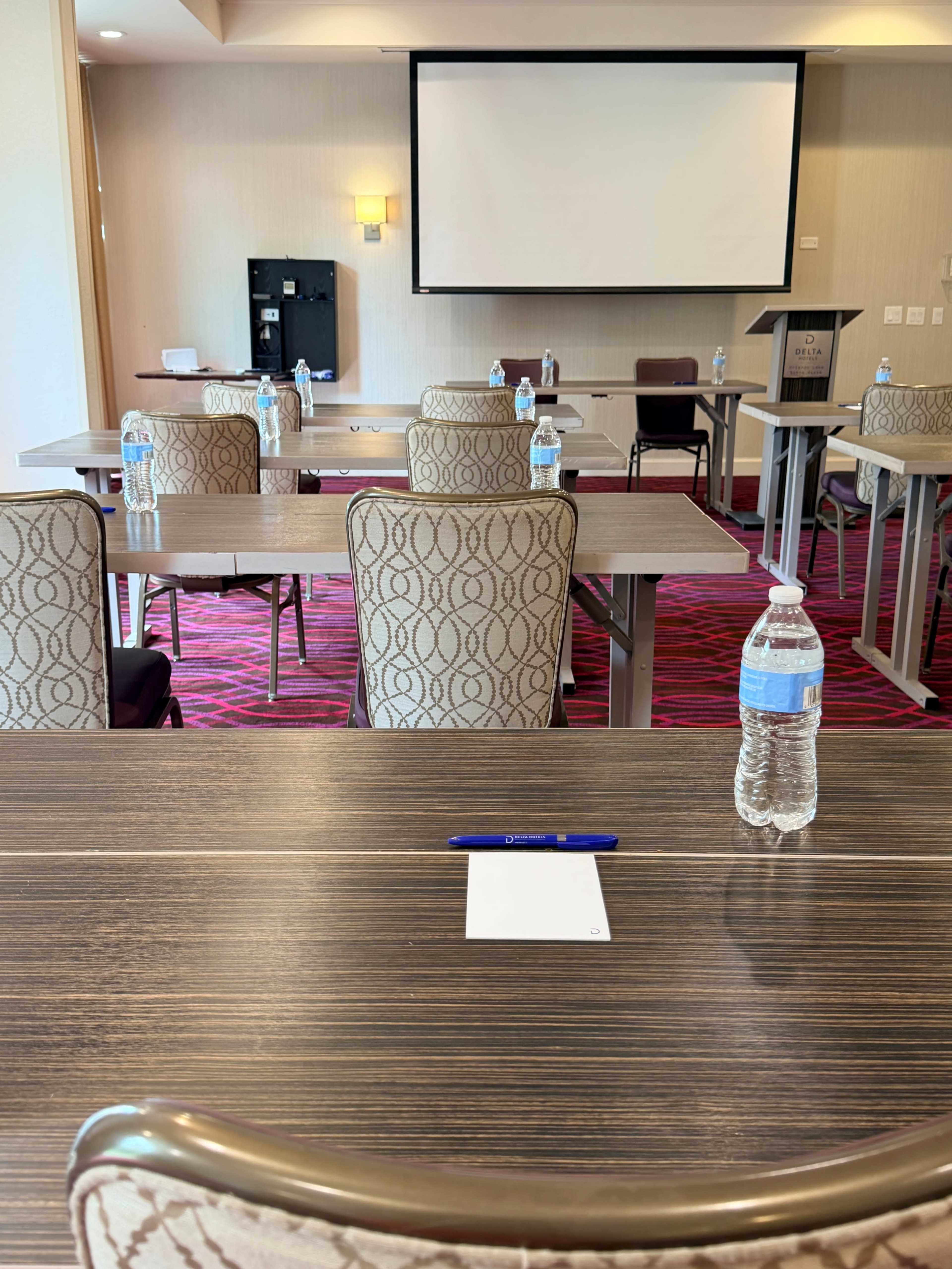 The image shows a conference room set up with several tables and chairs arranged in a row, equipped with water bottles and a notepad at each place setting, facing a presentation screen.