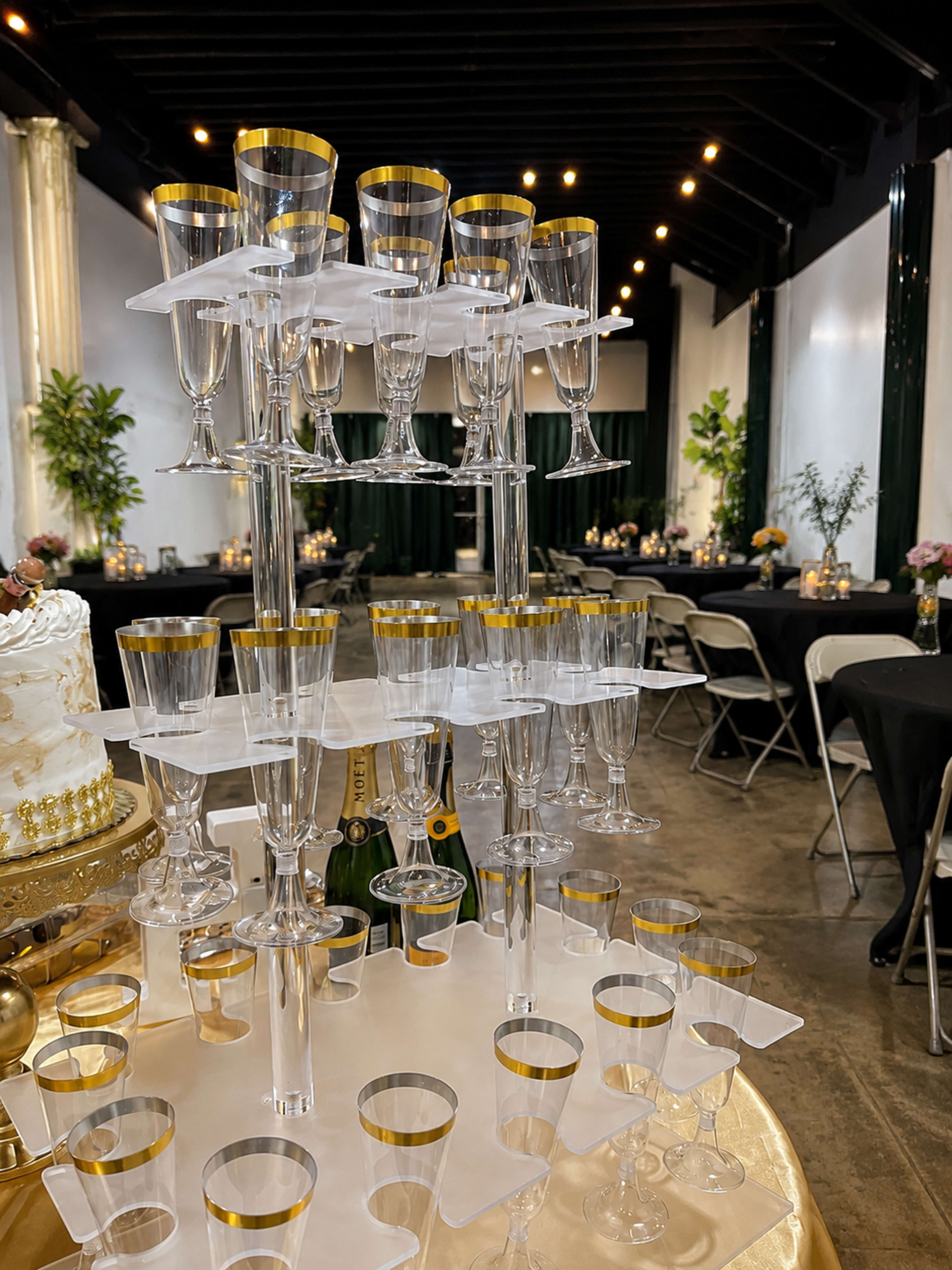 The image shows a tiered champagne glass display set up on a table, surrounded by decorated tables with floral arrangements and soft lighting in a banquet hall.