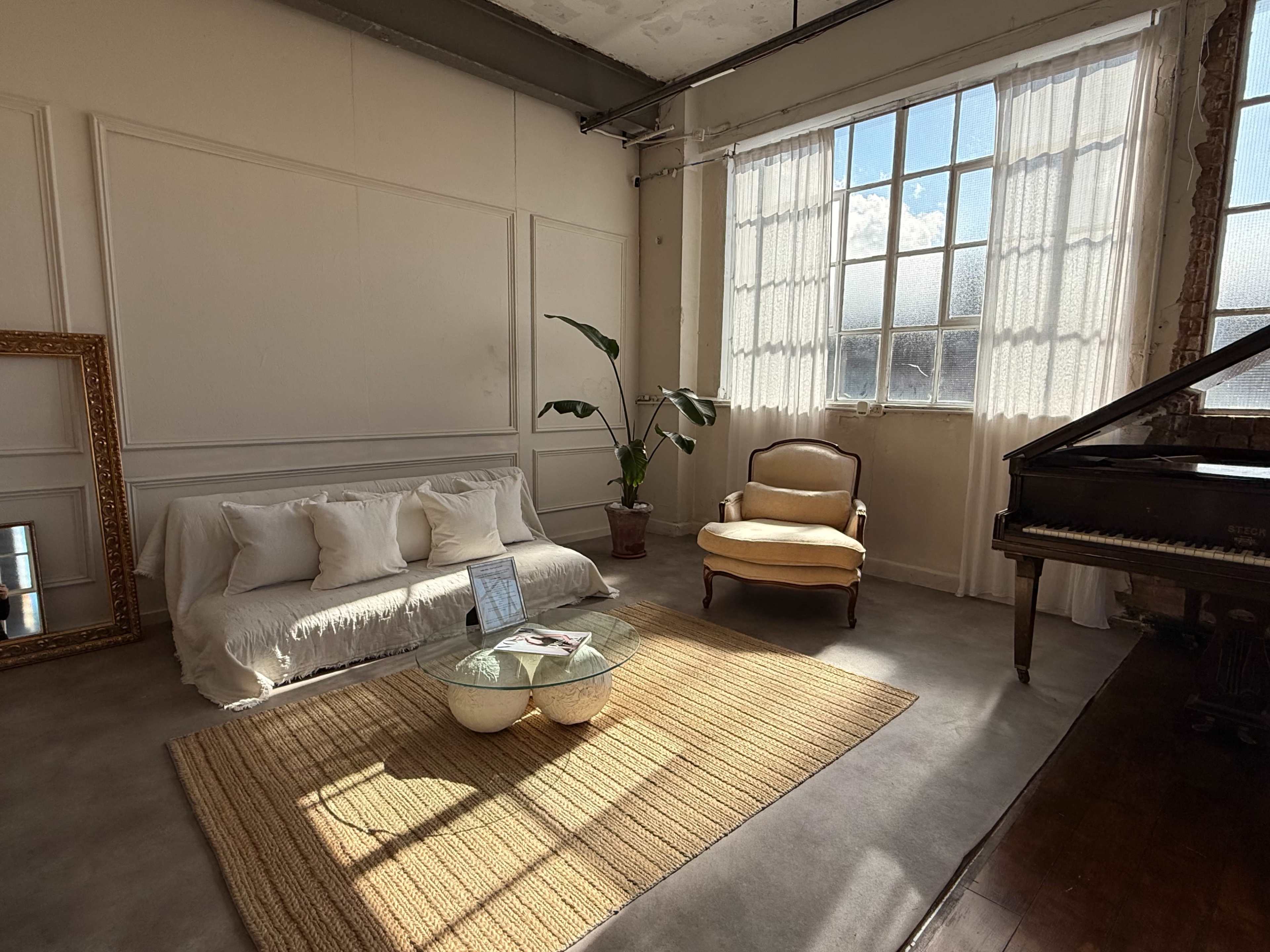 The image shows a bright, open room featuring a sofa with white cushions, an armchair, a grand piano, a glass coffee table, and a large window with sheer curtains.