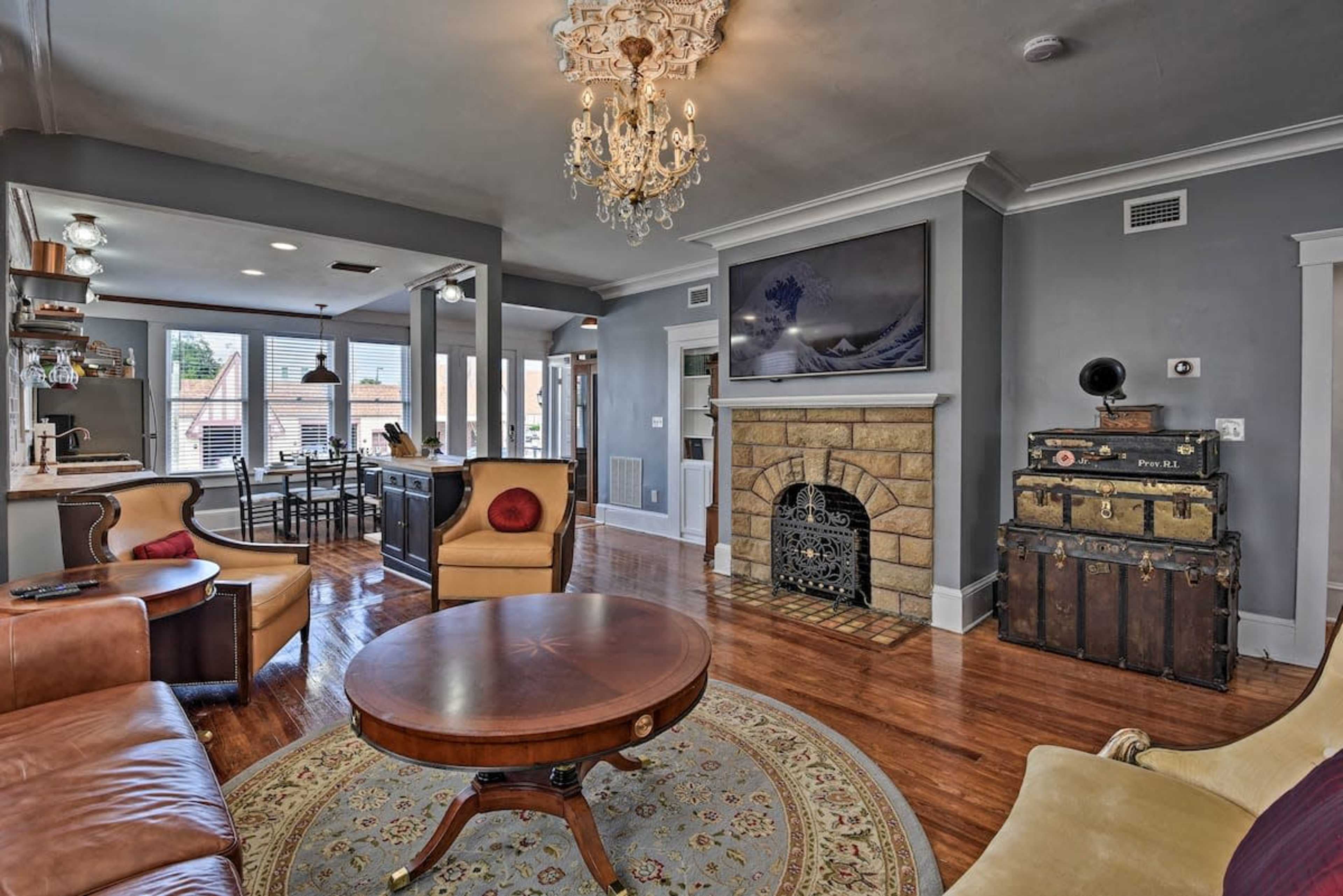 The image shows a living room featuring a chandelier, a circular rug, a vintage trunk, a stone fireplace, and large windows overlooking an outdoor view.