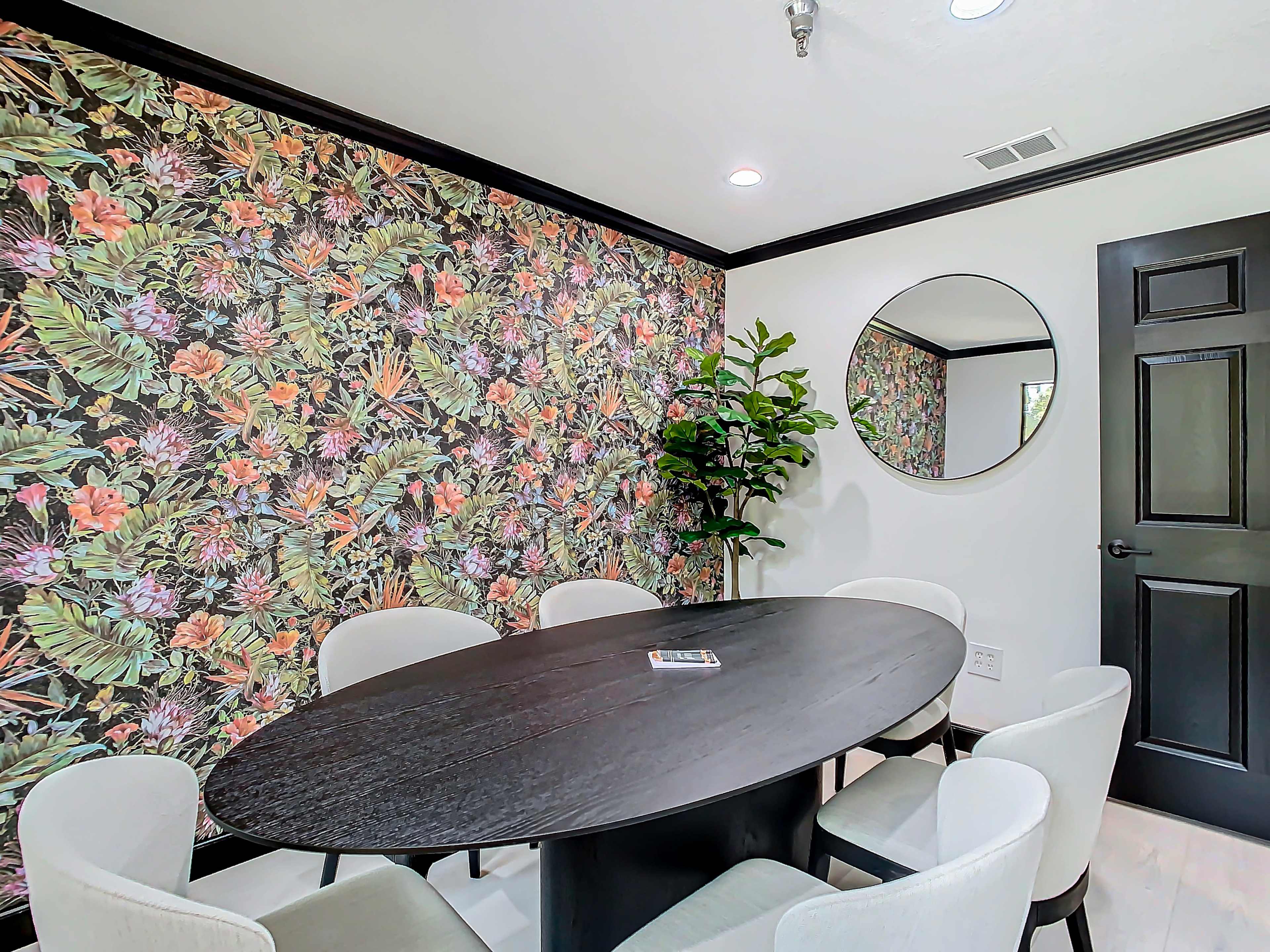 A round black dining table surrounded by white chairs is set against a floral wallpapered wall, with a mirror and a potted plant in the corner.