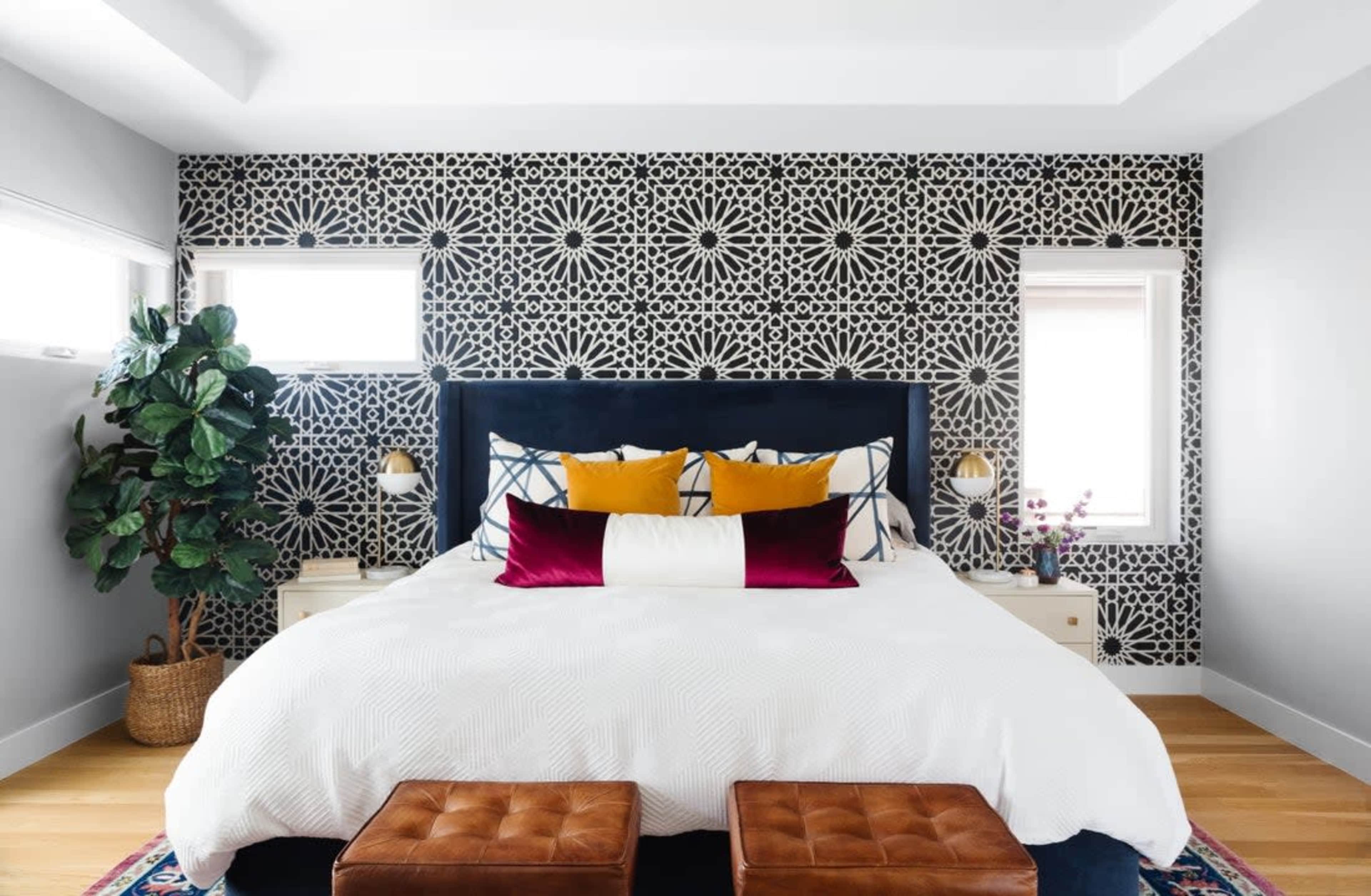 The image shows a modern bedroom featuring a large bed with a patterned black and white accent wall, flanked by two windows, and accompanied by a pair of brown ottomans at the foot of the bed.