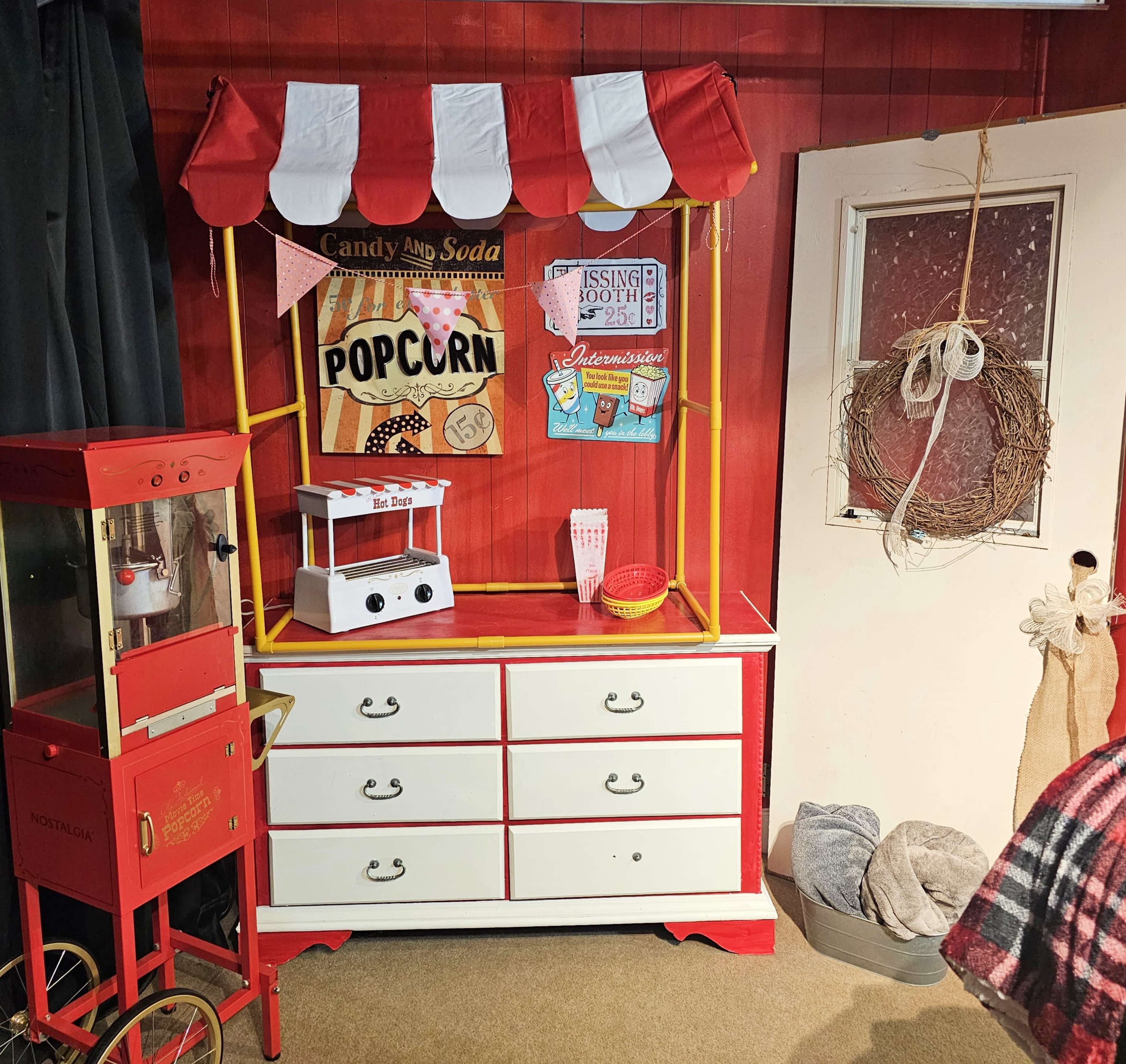 A vintage-style popcorn stand with a striped canopy, situated beside a red popcorn machine and decorated with nostalgic signs, against a red wall.