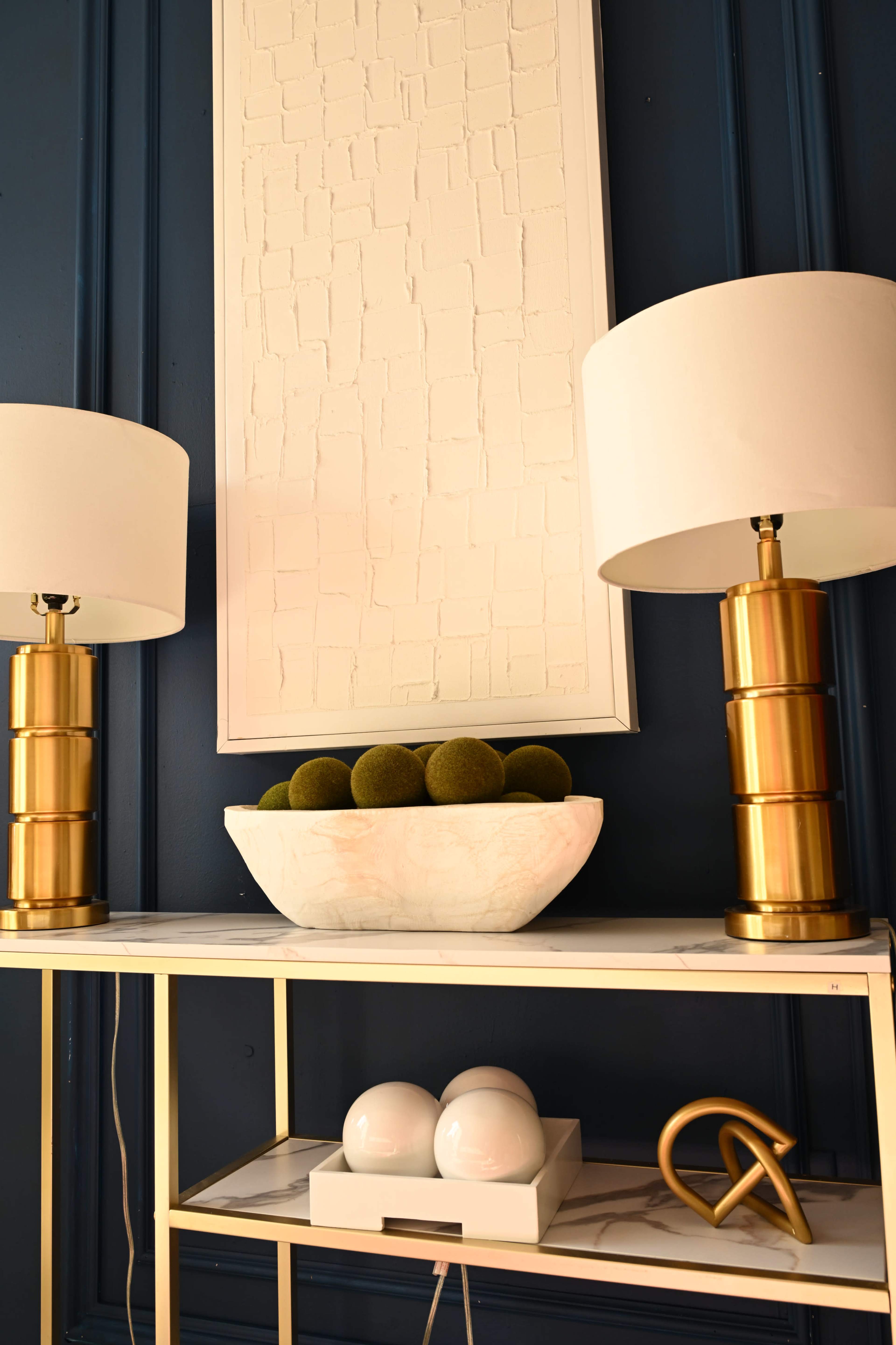 The image features a light-colored decorative bowl filled with green spheres, placed on a modern gold and white table, flanked by two gold lamps with white shades, against a dark blue wall.