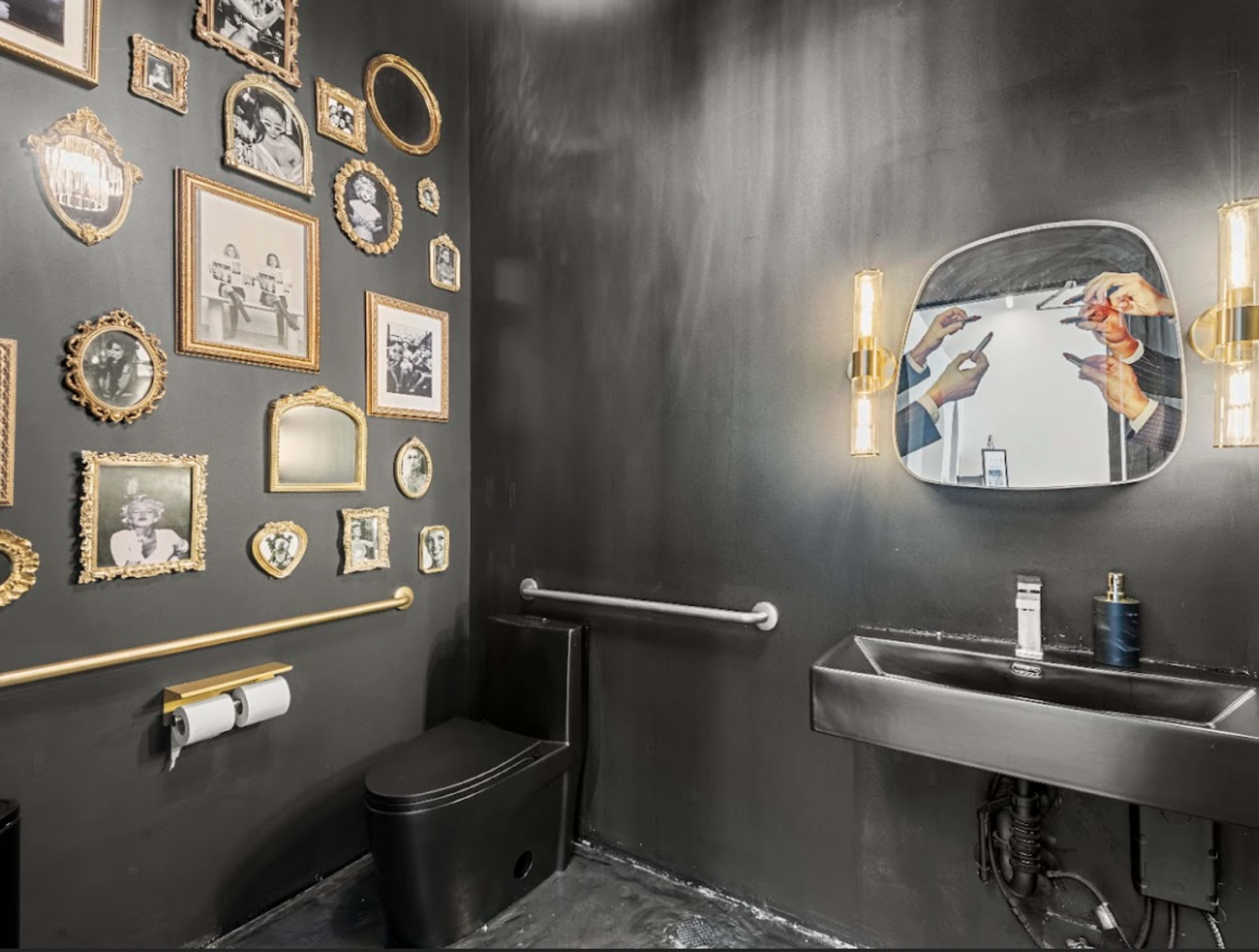 The image shows a modern bathroom with black walls, featuring a variety of framed pictures on one wall, and a sleek sink and toilet on the opposite side.
