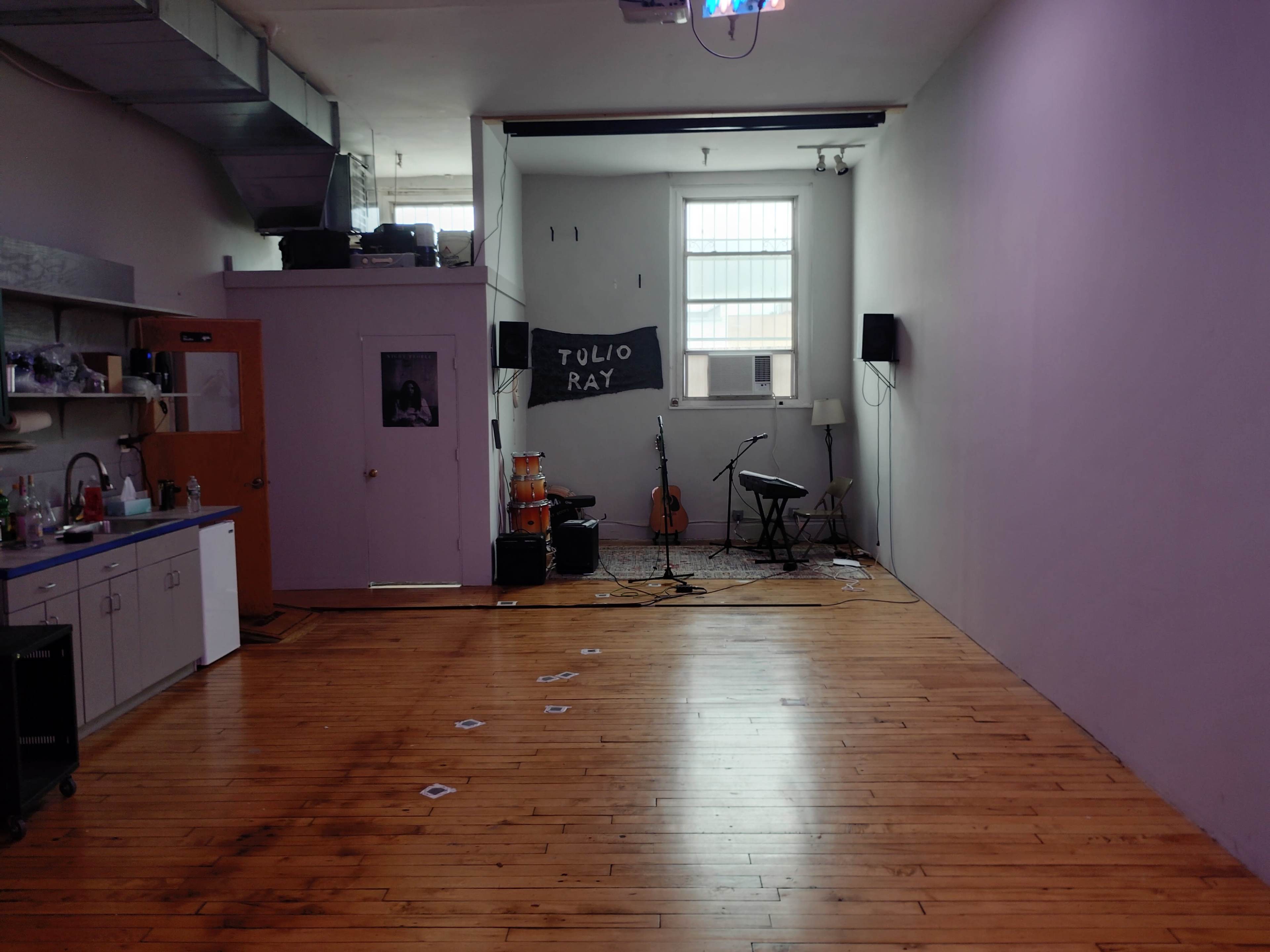 The image shows a spacious, empty room with wooden floors, a small kitchen area, a few musical instruments set up, and a chalkboard displaying the name "TULIO RAY" on the wall.