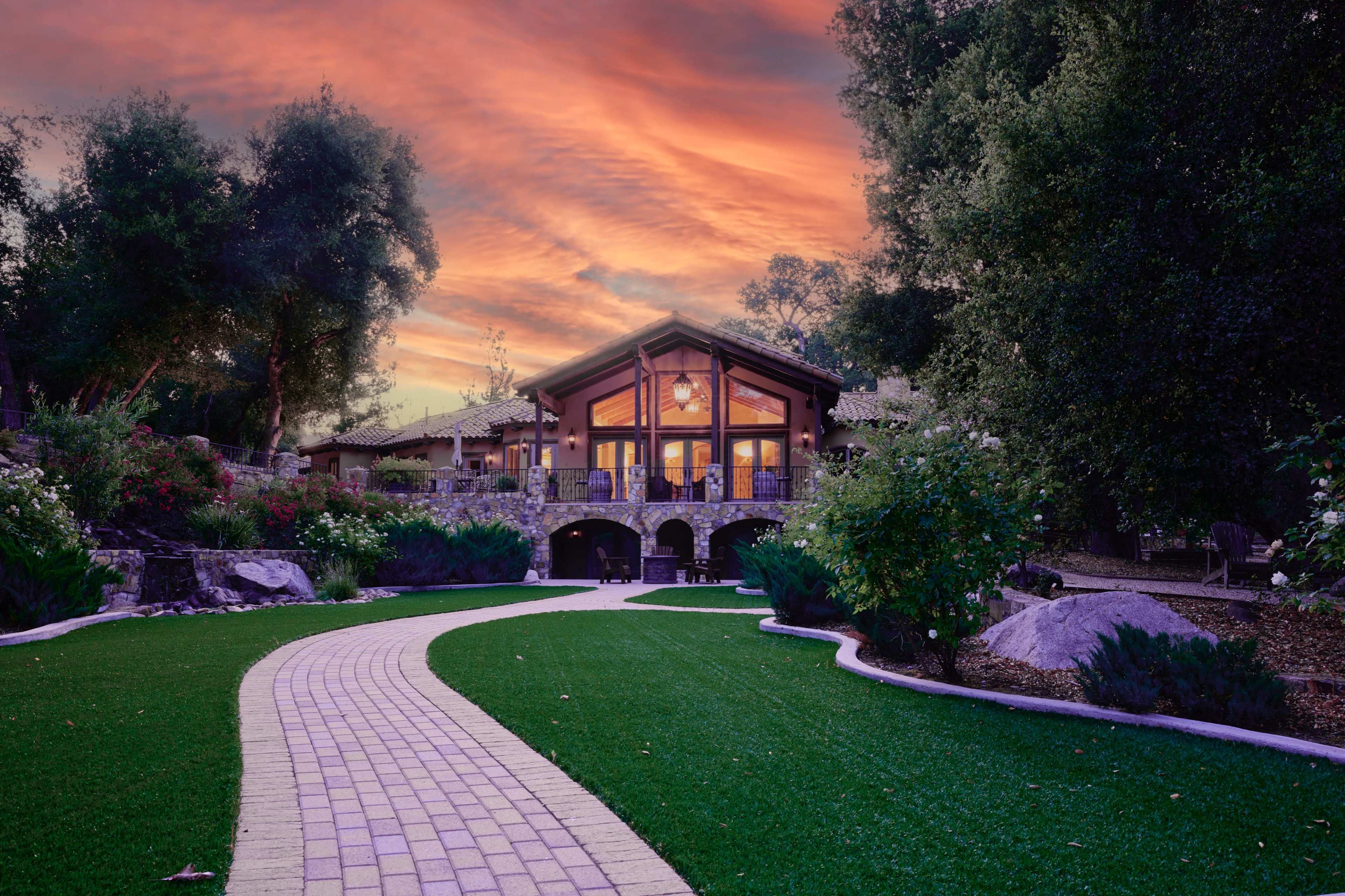 A stone pathway leads to a modern house surrounded by lush greenery and blooming flowers under a colorful sunset.