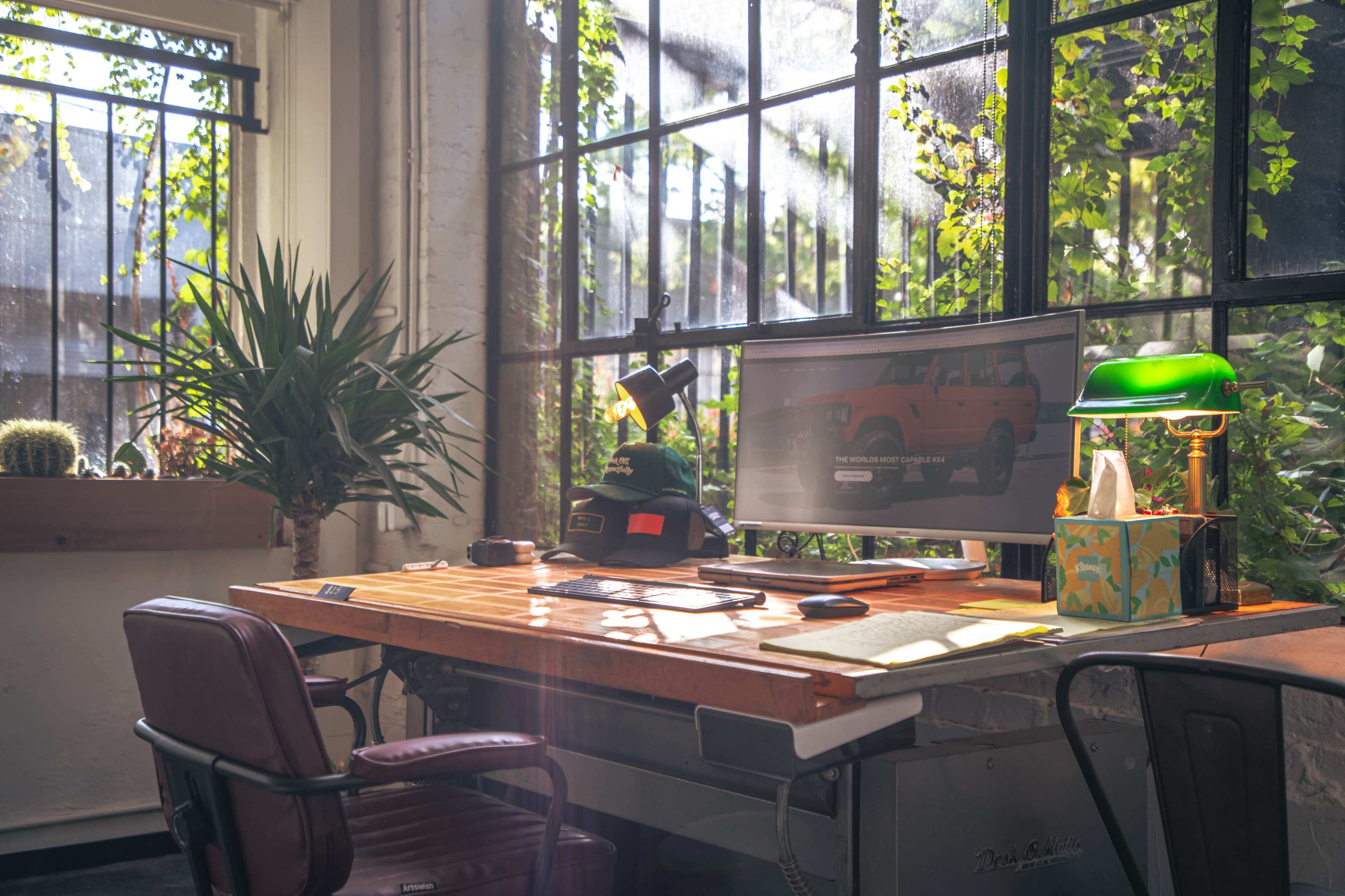 A wooden desk with a computer monitor, lamp, and stationery is positioned in a brightly lit room surrounded by large windows and plants.