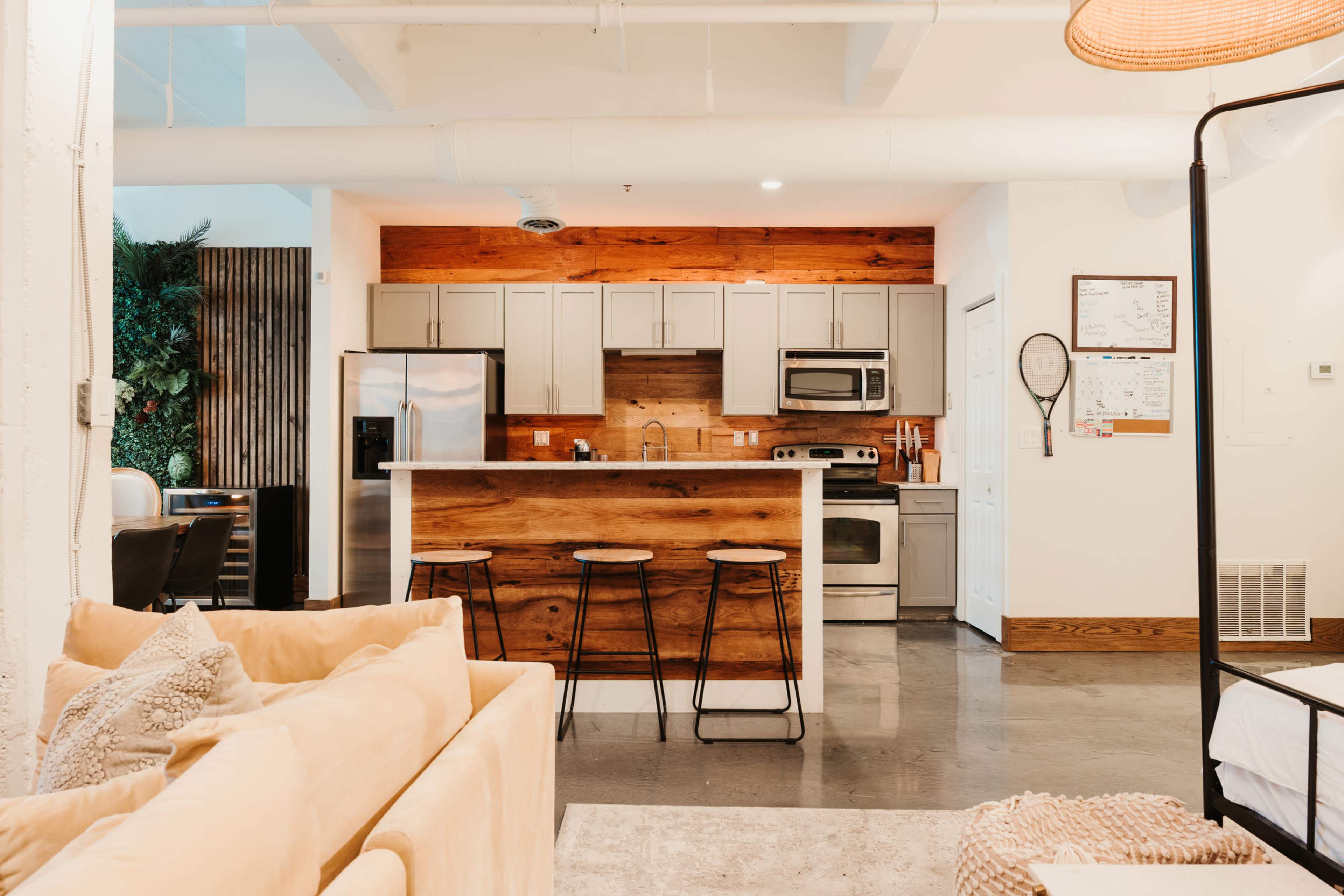The image shows a modern kitchen with a wooden accent wall, stainless steel appliances, and three bar stools facing a counter.