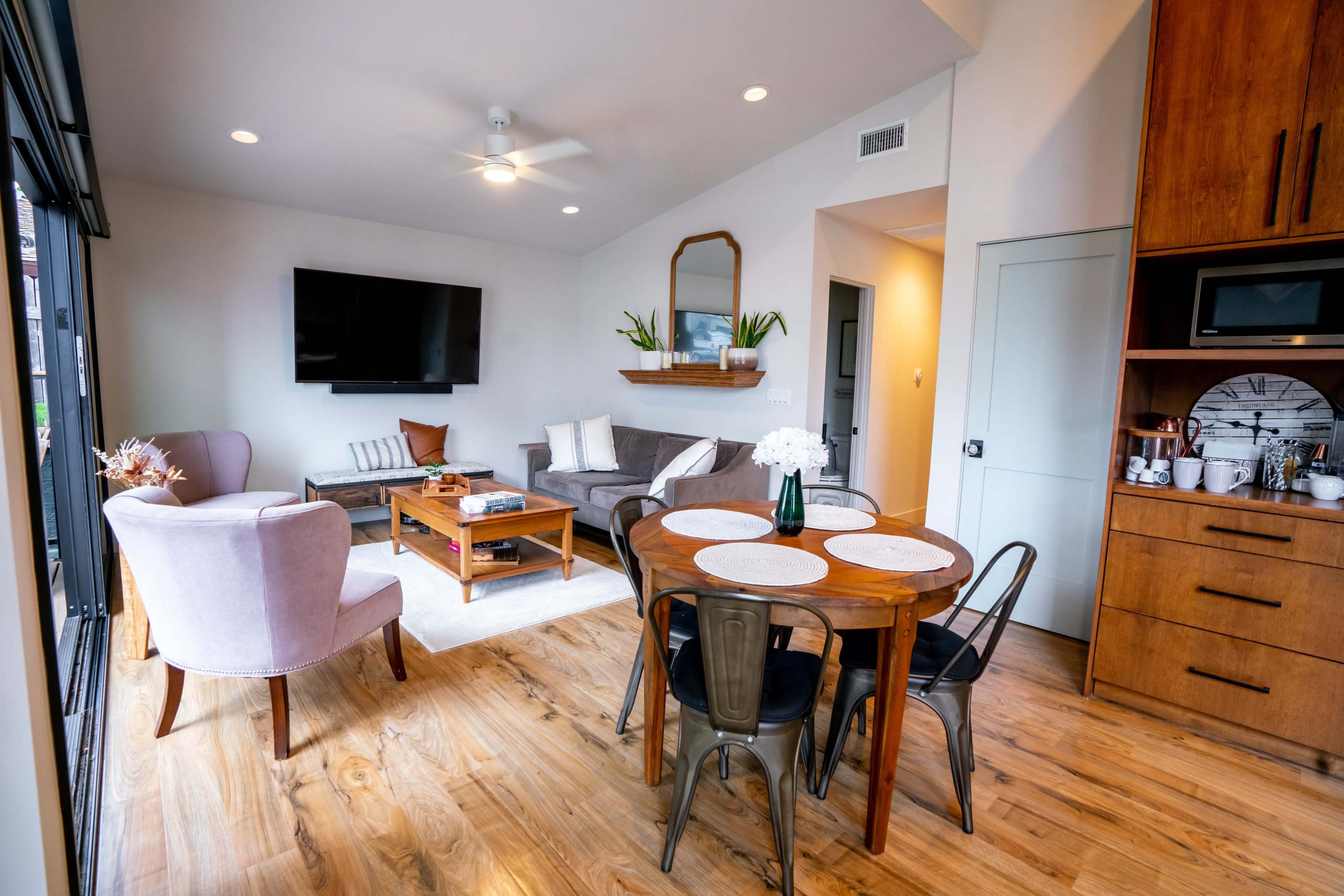 A modern living space features a seating area with a sofa, two chairs, a coffee table, and a dining table surrounded by metal chairs, with natural light coming from large sliding doors.