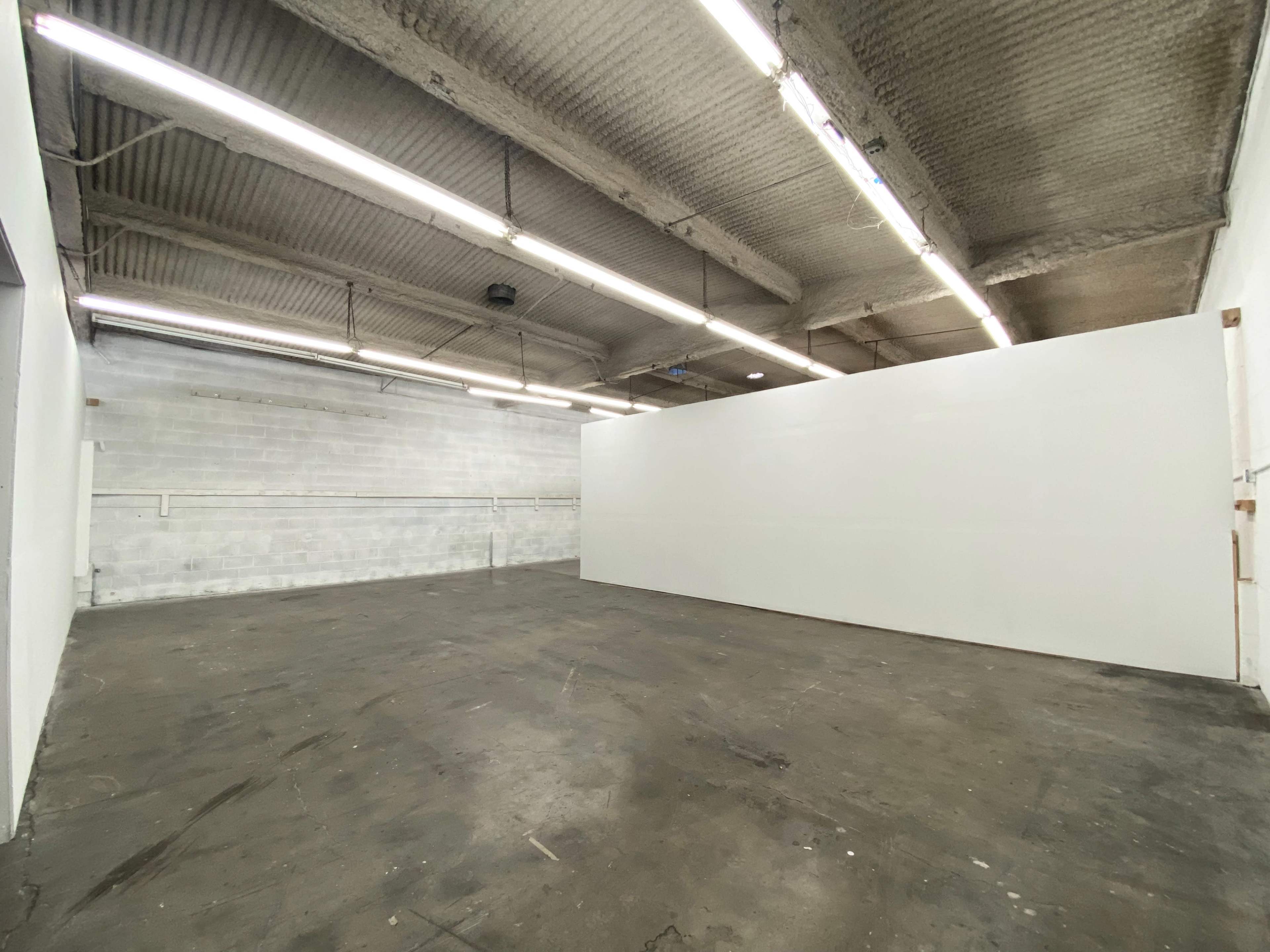An empty industrial space with white walls, a concrete floor, and overhead fluorescent lighting.
