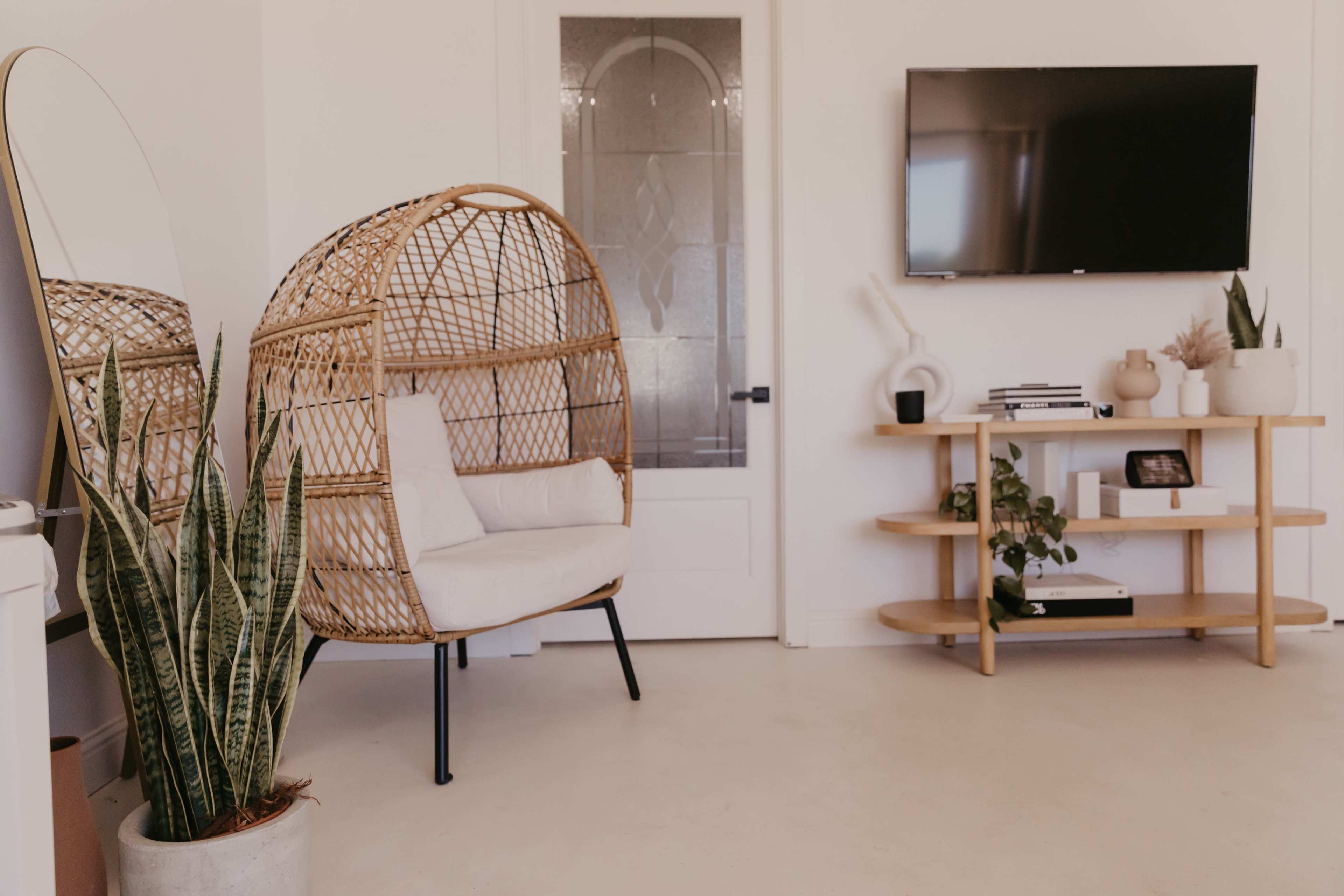 A modern room featuring a rattan chair, a tall plant, a wall-mounted television, and a small wooden shelf with various decorative items.