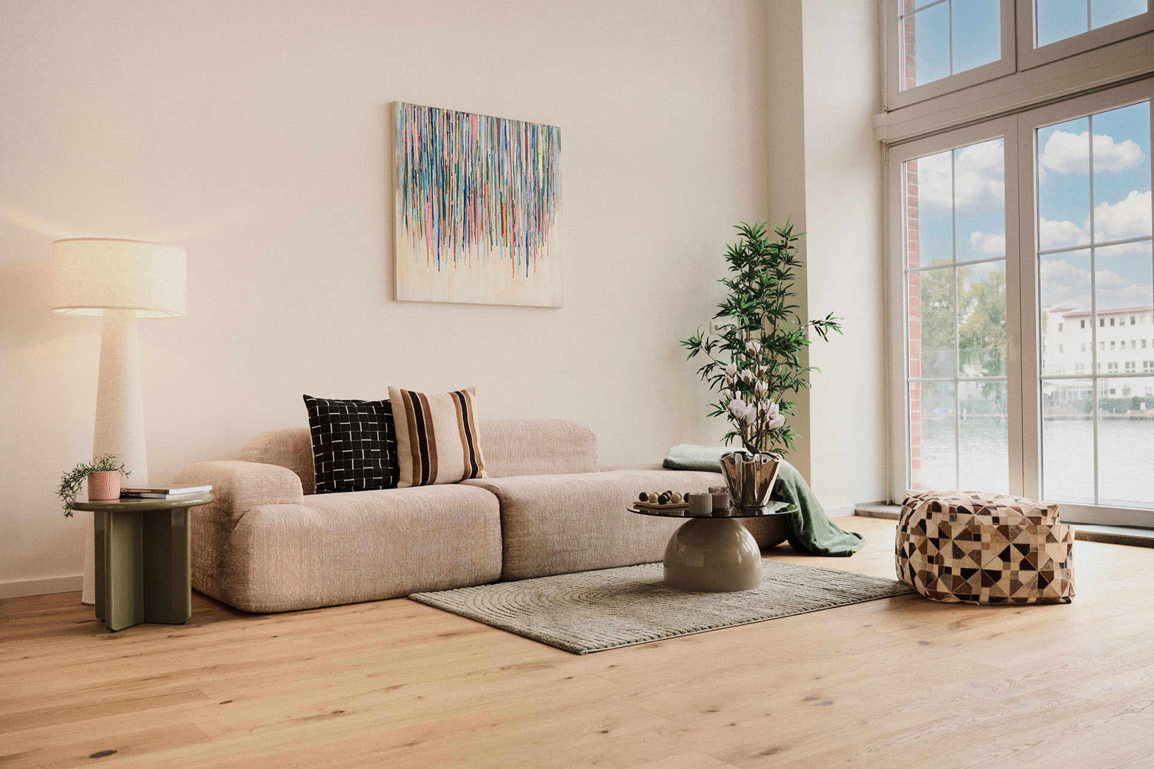 The image features a modern living room with a light-colored sofa, a decorative lamp, a small table, and large windows showcasing a view of the outdoors.