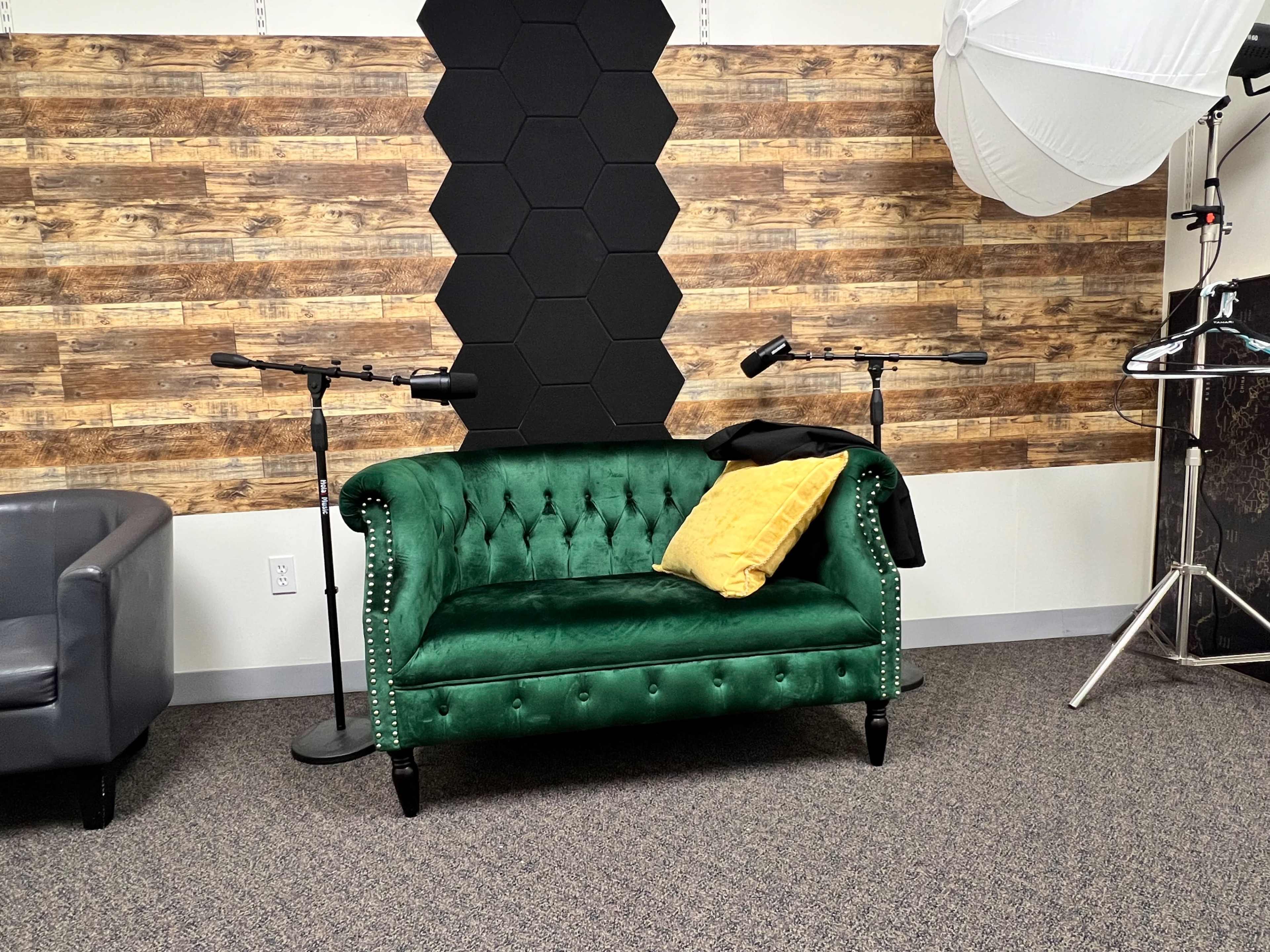 A green tufted sofa with a yellow pillow sits against a backdrop of textured wood panels and lighting equipment in a studio setting.