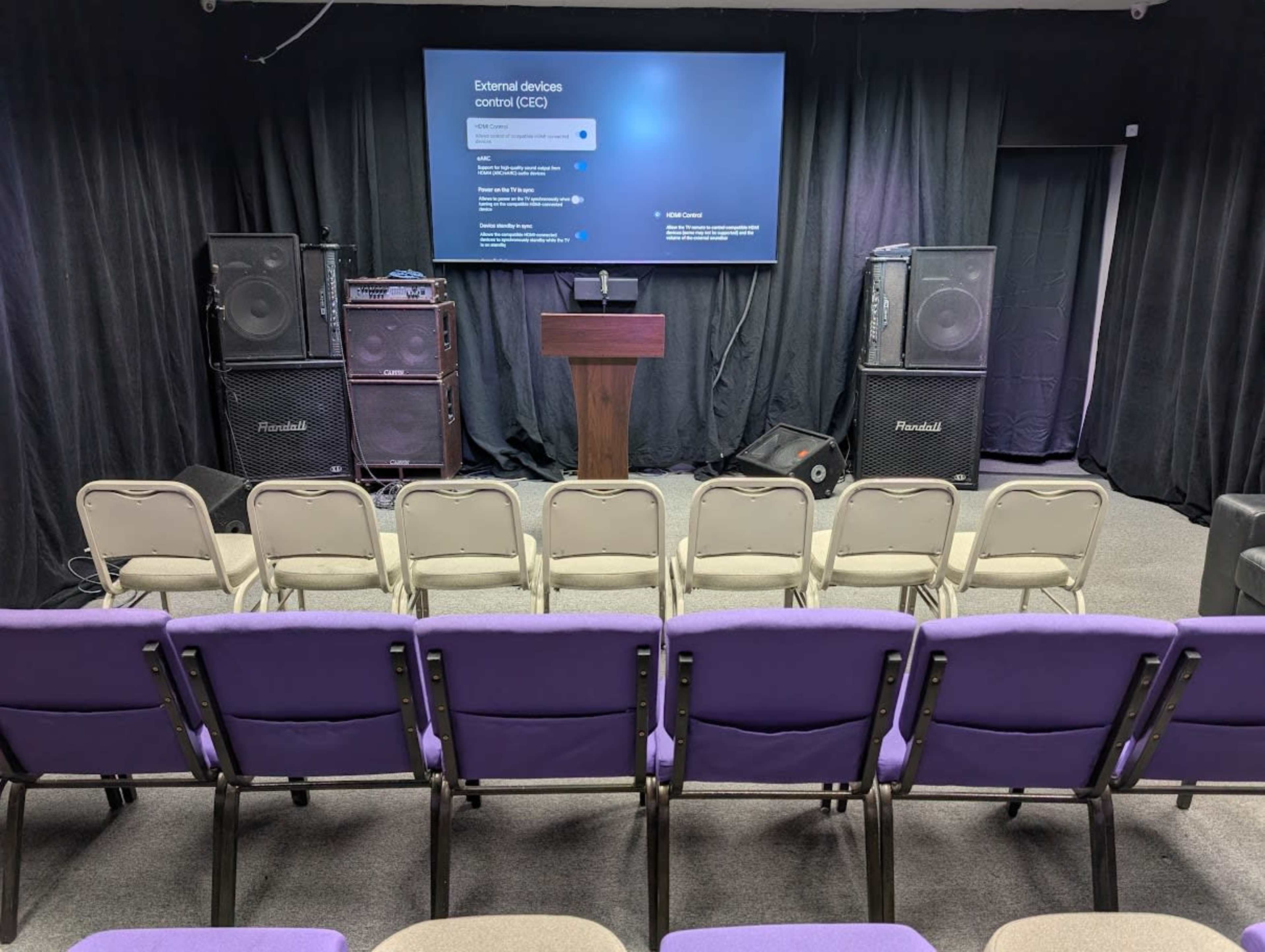 The image shows a small auditorium setup with rows of purple chairs facing a podium and a large screen, flanked by two speaker systems.