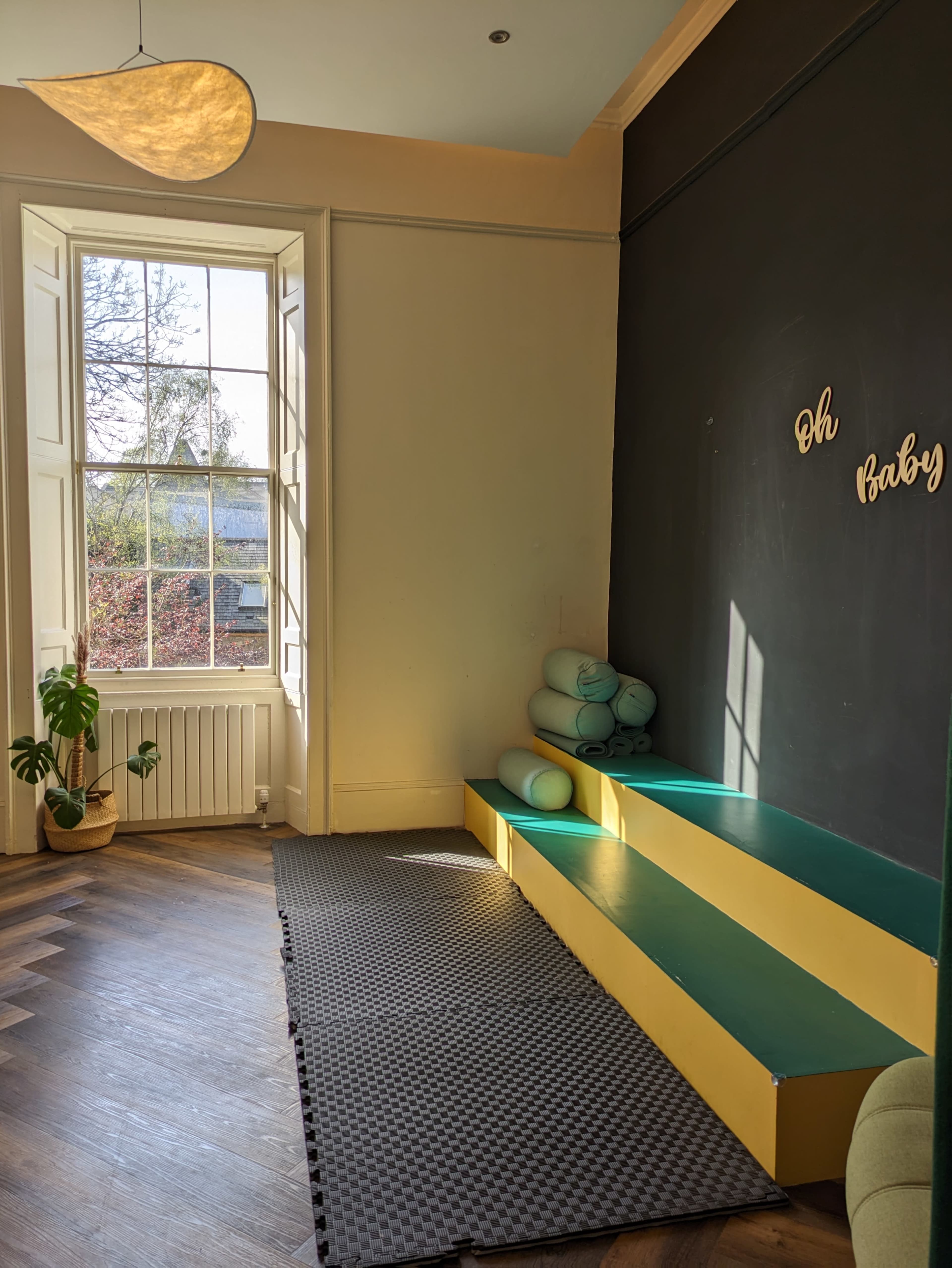 A bright room with large windows, a yellow step area, and stacked exercise mats along a dark green wall with the phrase "Oh Baby" displayed.