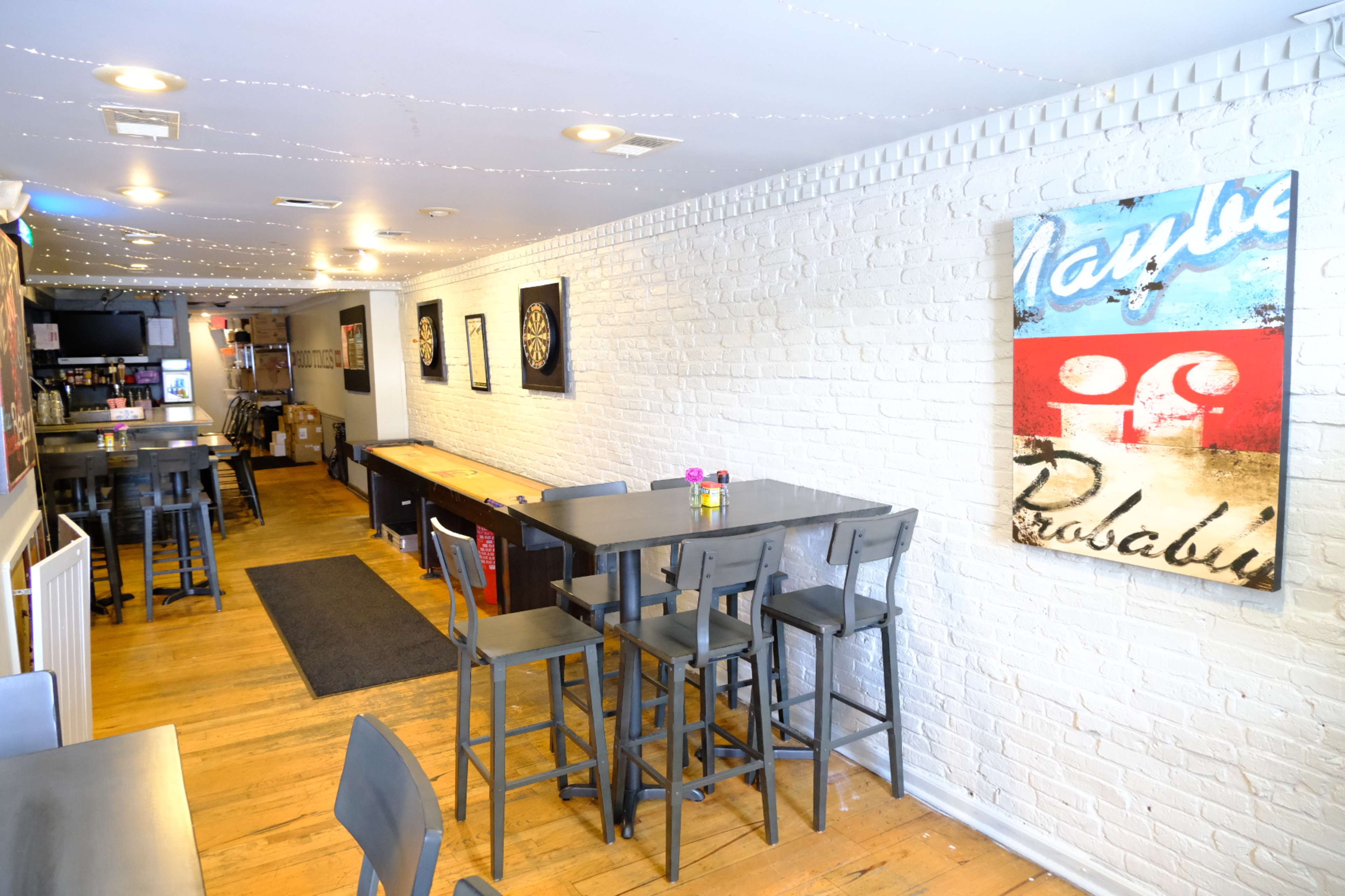 A narrow, well-lit bar area with wooden flooring, high tables, and chairs, alongside a white brick wall adorned with artwork.