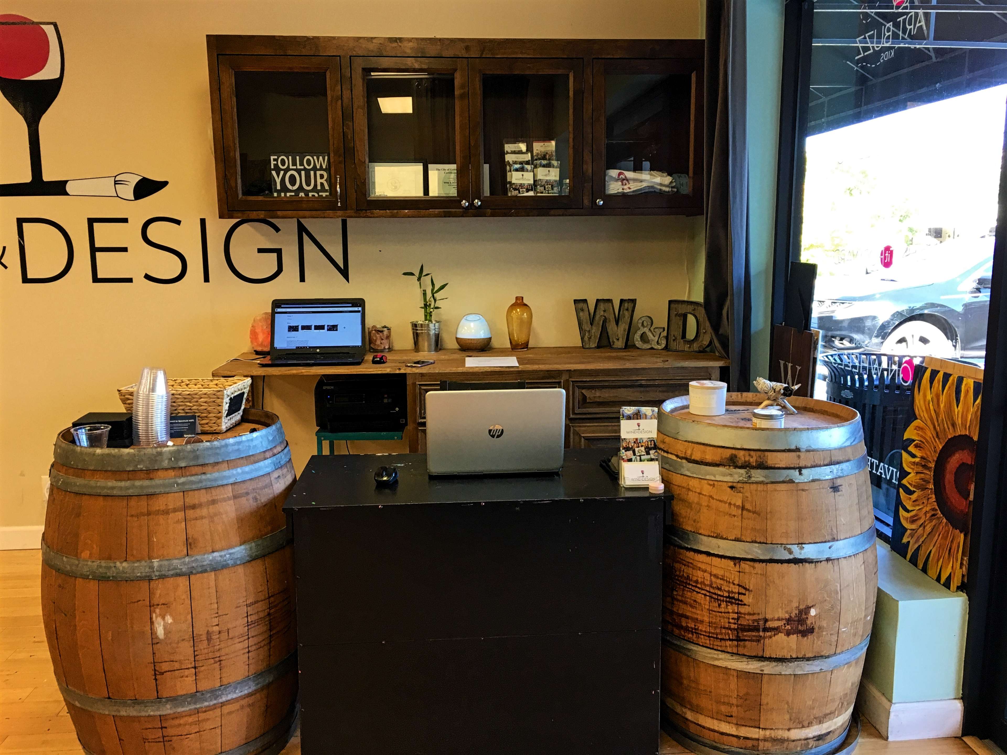 A reception area featuring two wooden barrels flanking a black desk with a laptop and various office supplies, against a wall with a wine glass graphic and a sign that reads "FOLLOW YOUR LEAD."
