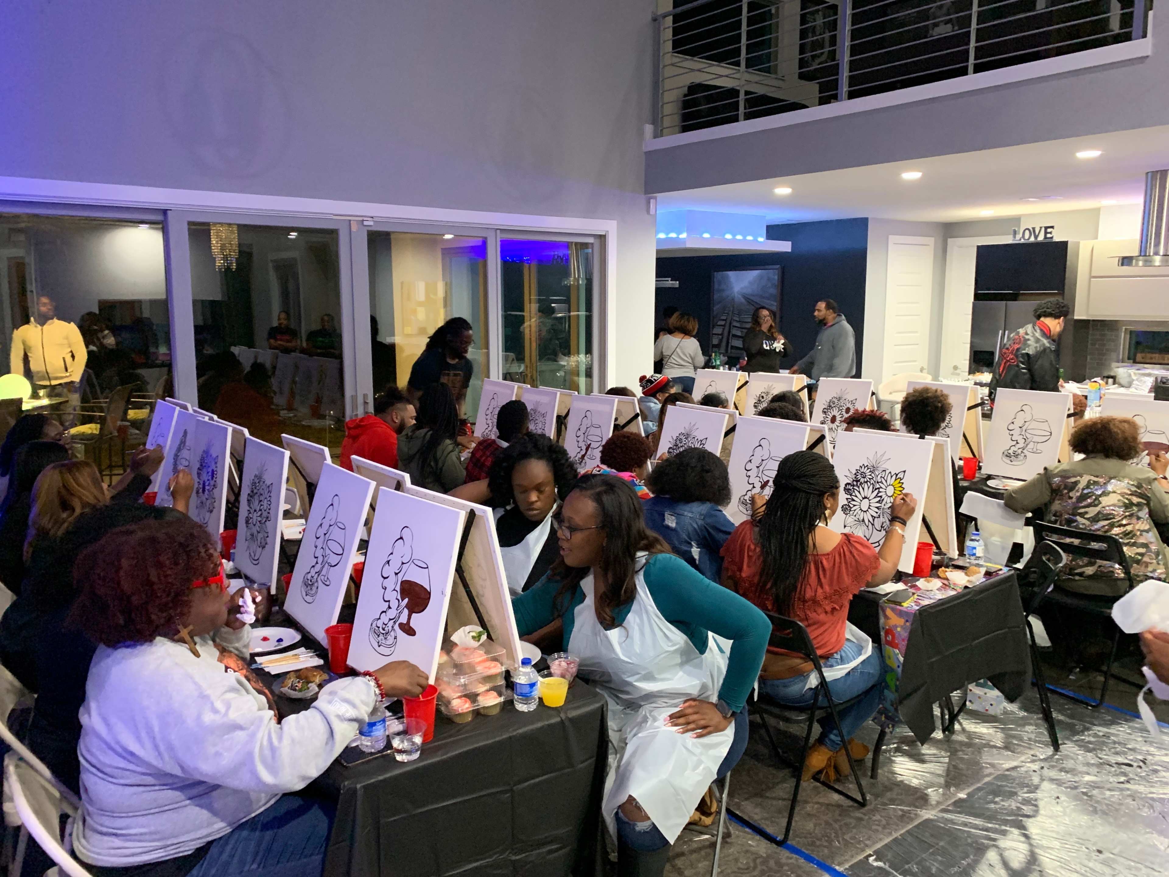 A group of people participate in a painting class, each working on canvas in a well-lit indoor space.