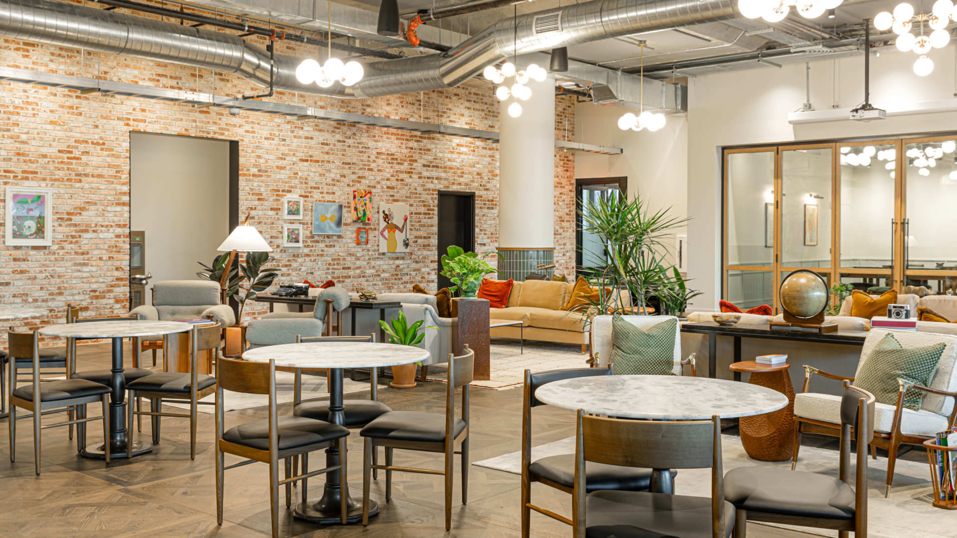 A modern co-working space with brick walls, circular tables, and comfortable seating areas surrounded by plants and decorative elements.