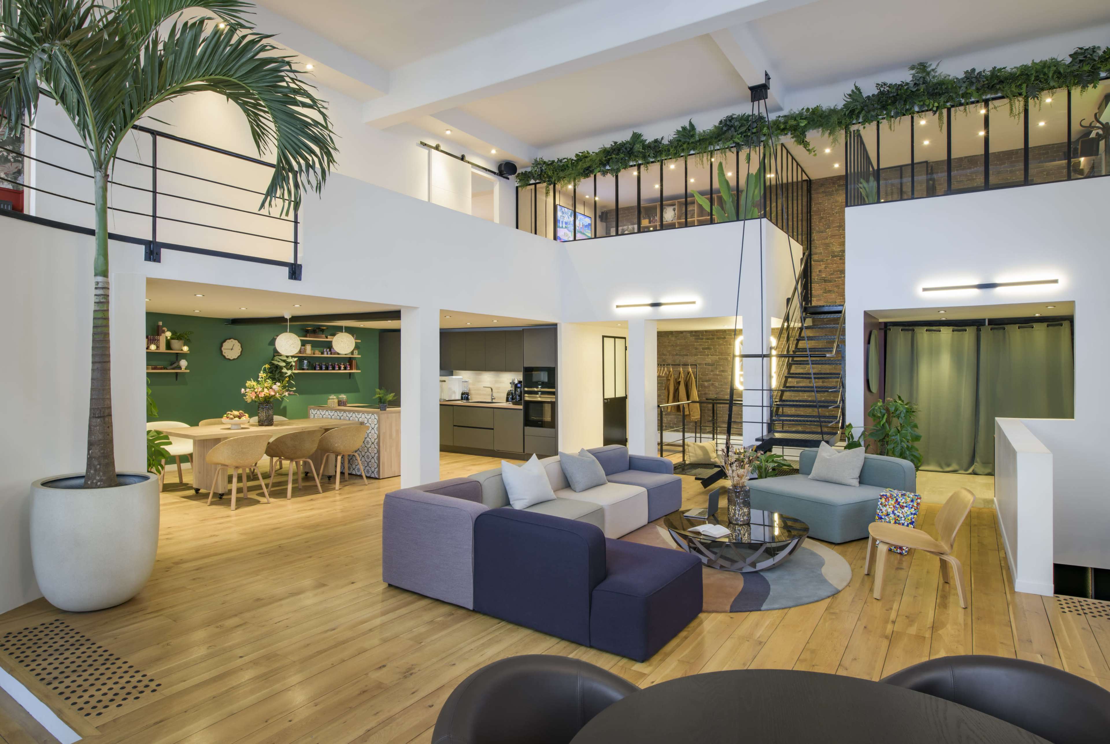 The image shows a modern, open-concept living space with a multi-level design, featuring a lounge area, dining area, and a staircase.