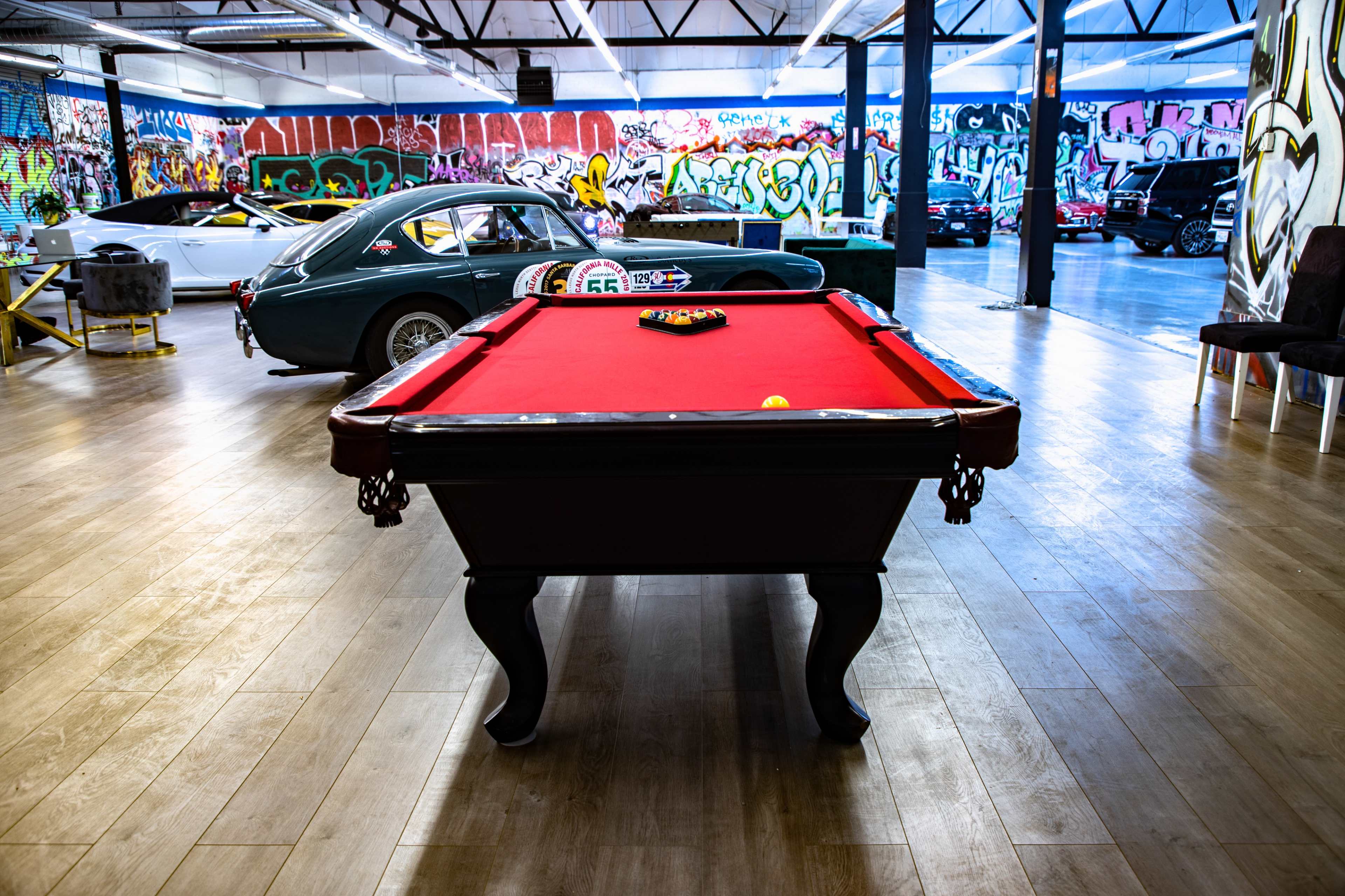 A red pool table is centered in a spacious garage with colorful graffiti on the walls and classic cars nearby.