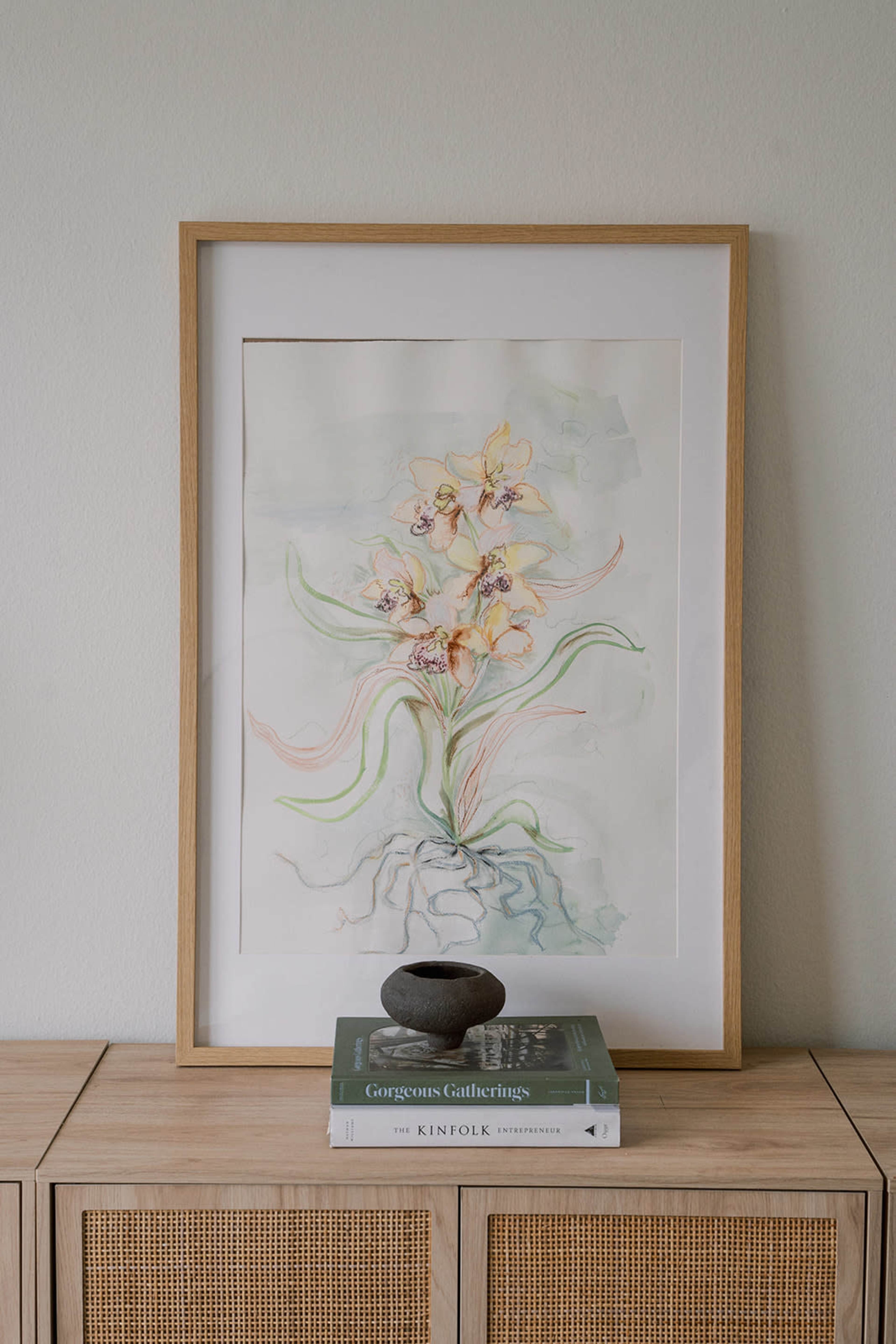A framed watercolor painting of yellow flowers is displayed above a stack of books on a wooden surface.