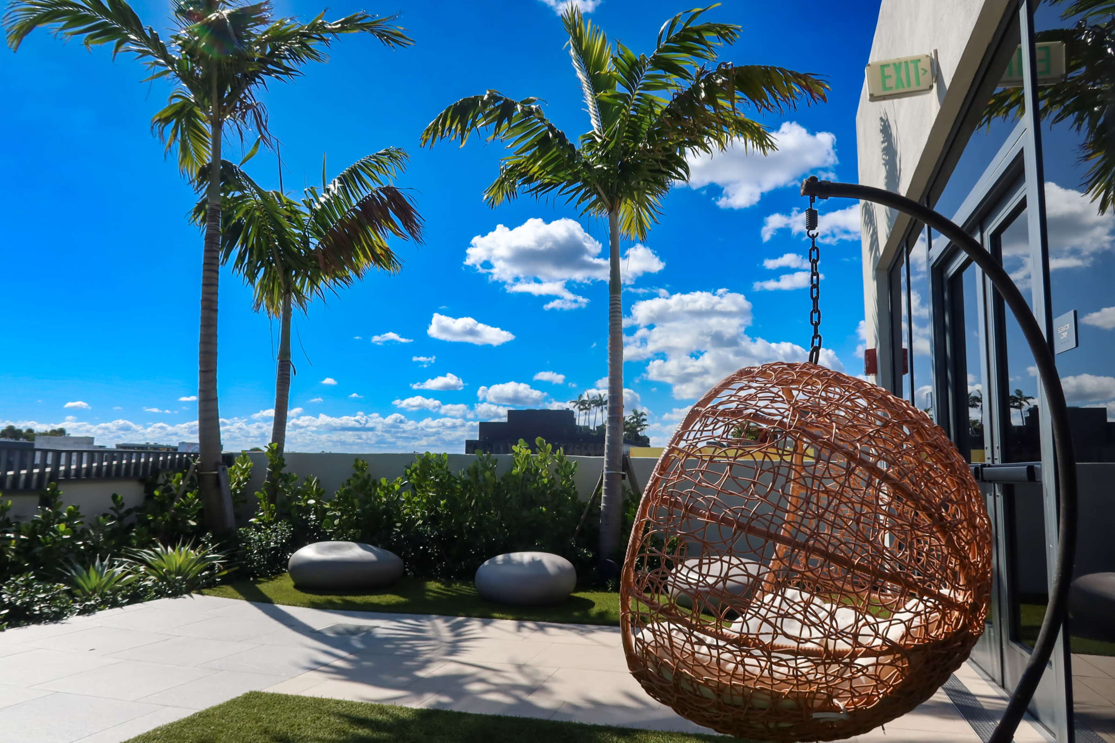 A hanging basket chair is positioned outdoors among palm trees under a bright blue sky with fluffy clouds.