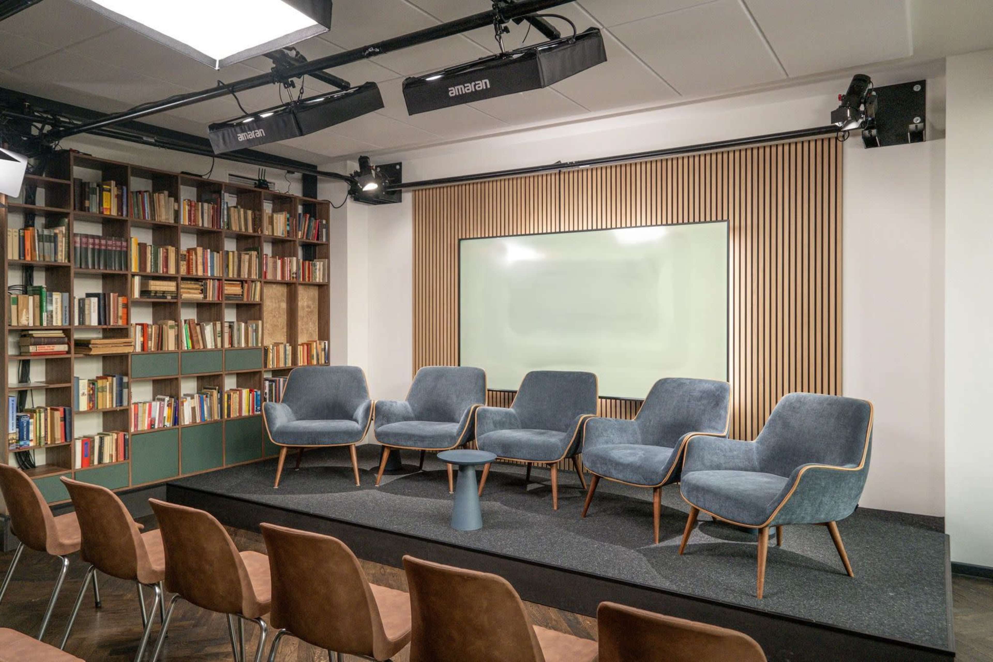 The image shows a modern presentation space with five blue upholstered chairs arranged in a semicircle around a small table, situated in front of a large screen and a backdrop of bookshelves filled with books.