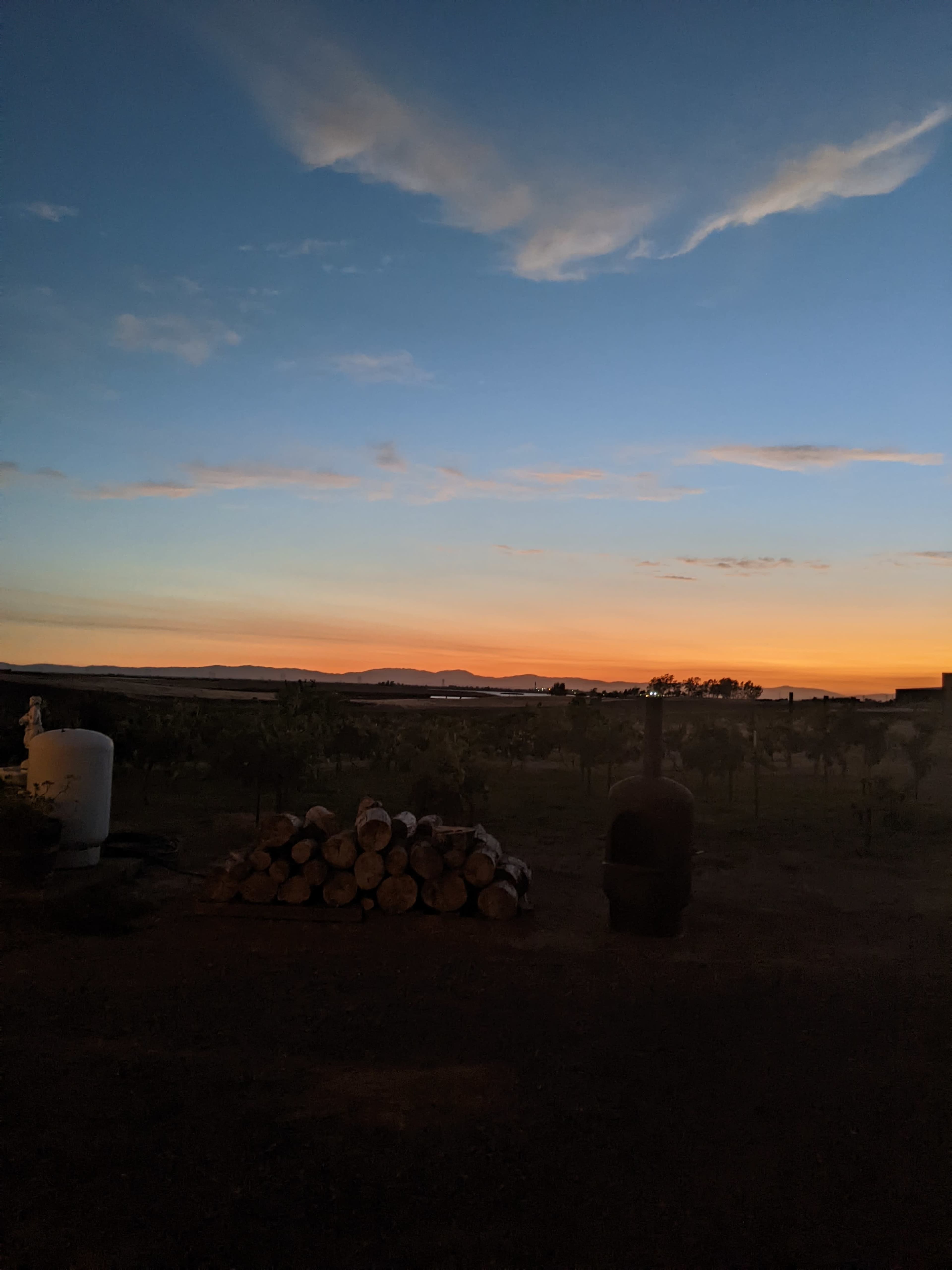 A rural landscape at sunset with stacked logs in the foreground and a gradient sky transitioning from orange to blue.